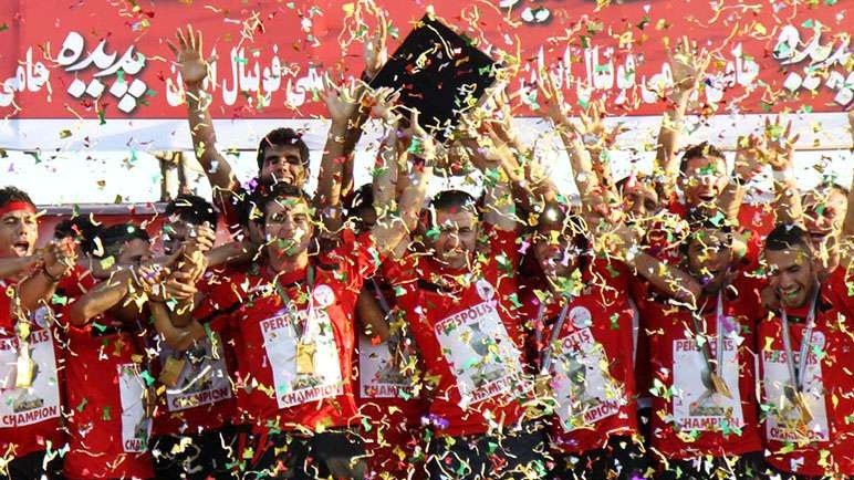 L'image montre une équipe de football célébrant une victoire. Les membres de l'équipe portent des maillots rouges et affichent des sourires de joie. Ils tiennent des trophées et sont entourés de confettis colorés qui tombent sur eux. L'ambiance est festive, et on peut sentir l'excitation de la victoire à travers leurs expressions. Des banderoles et des affiches derrière eux montrent le nom de l'équipe et le mot "champion".