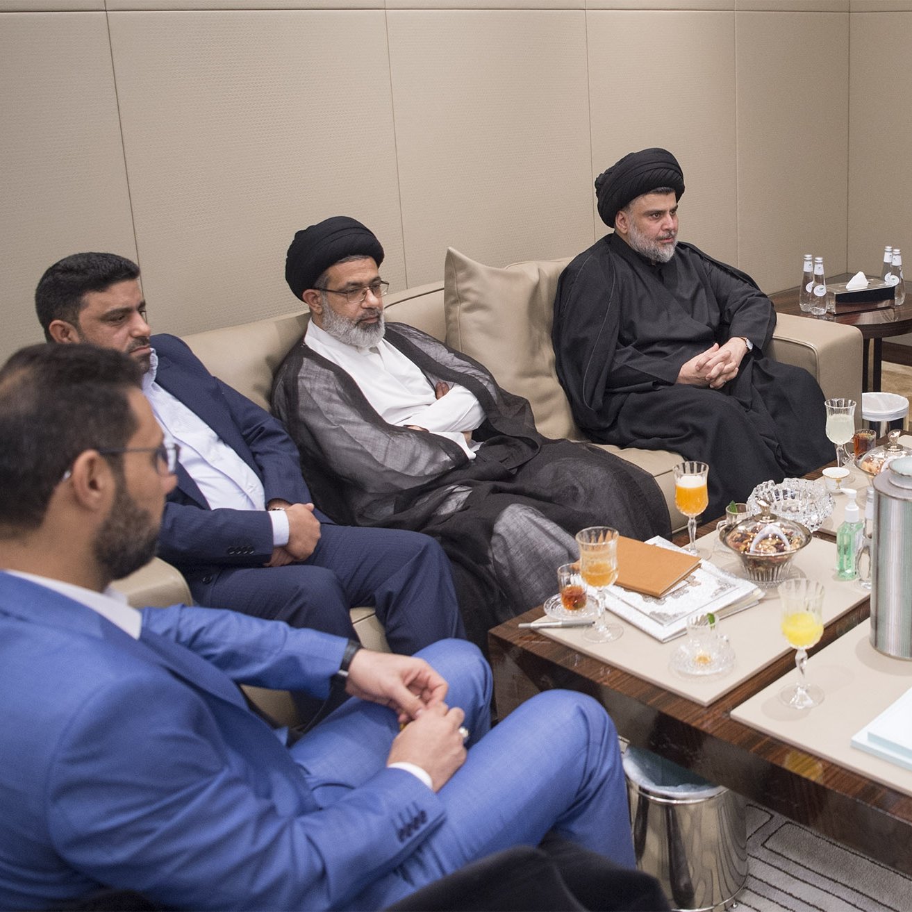 تظهر الصورة مجموعة من الأشخاص يجلسون في قاعة اجتماعات. يتواجد بعضهم بملابس تقليدية، بينما يرتدي آخرون ملابس رسمية. على الطاولة أمامهم توجد بعض المشروبات والوجبات الخفيفة. يبدو أن الاجتماع يتضمن مناقشات بين الأفراد، حيث يمتلك كل منهم تعبيرات جدية. المكان يبدو أنيقًا وعصريًا، مما يوحي بأهمية اللقاء.