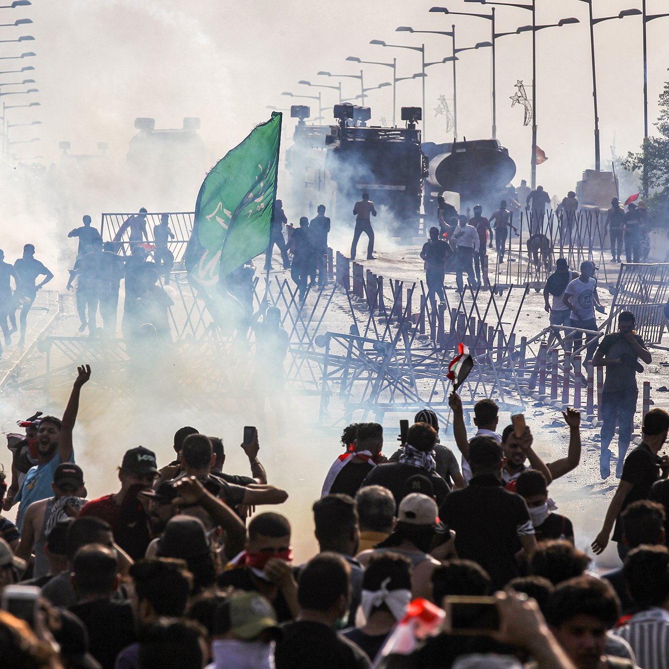 تصویر نشان‌دهنده یک تظاهرات یا اعتراض عمومی است که در یک خیابان یا پل در حال برگزاری است. تعداد زیادی از معترضان در حال حرکت و تجمع هستند و برخی از آن‌ها پرچم‌هایی را در دست دارند. دود ناشی از گاز اشک‌آور یا مواد دیگر در هوا دیده می‌شود و با ماشین‌های مخصوص پلیس در پس‌زمینه، فضایی تنش‌آلود به وجود آمده است. نوارهای موانعی نیز در اطراف معترضان مشاهده می‌شود که برای کنترل جمعیت و جلوگیری از پیشروی آن‌ها استفاده می‌شود.
