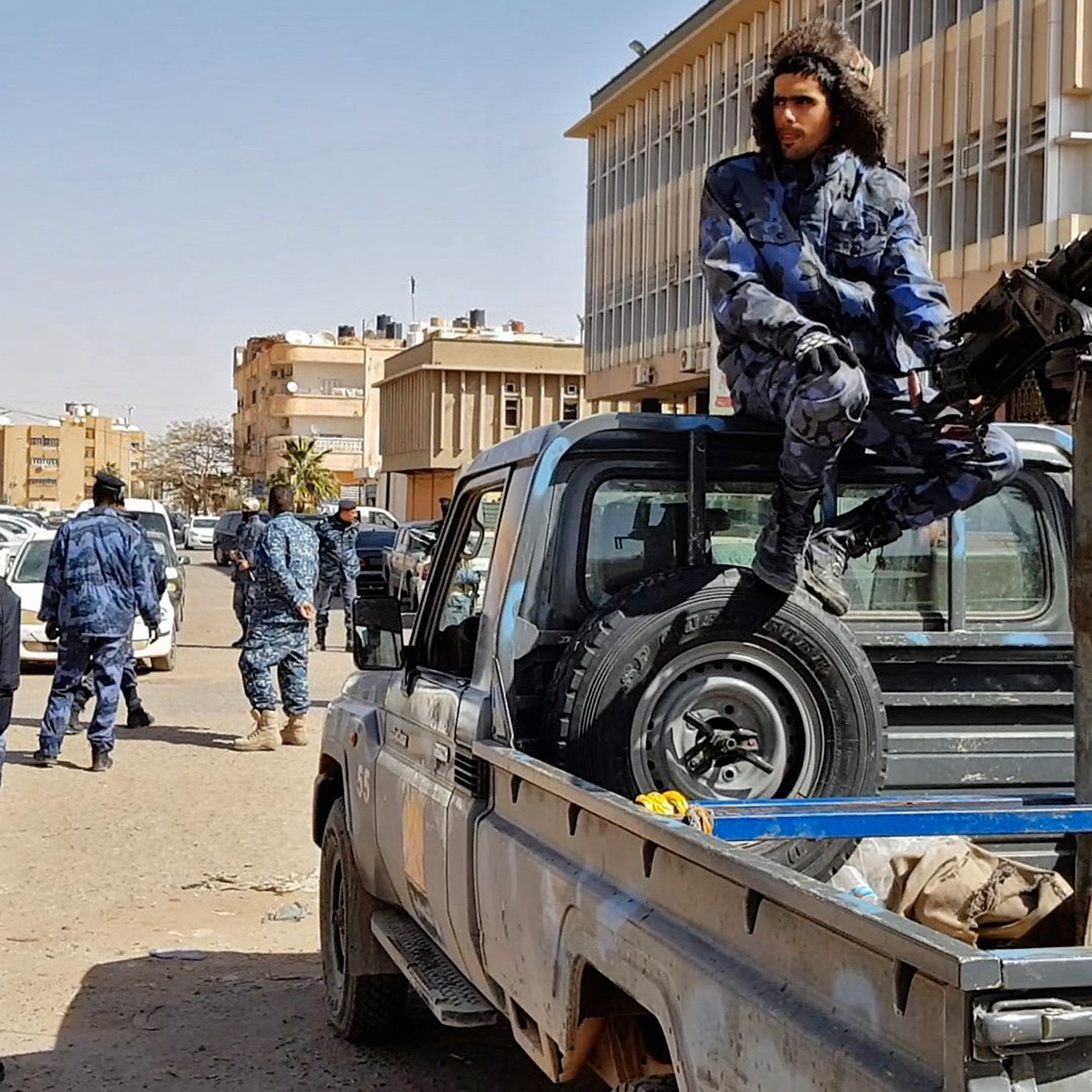 L'image montre une scène urbaine impliquant des forces de sécurité. On peut y voir plusieurs policiers en uniforme, certains armés, se déplaçant autour de véhicules. Un homme est assis sur le plateau d'une camionnette, près d'une arme lourde montée sur le véhicule. L'environnement semble être une ville moderne, avec des bâtiments en arrière-plan. L'ambiance générale de l'image suggère une opération de sécurité ou une situation de contrôle.