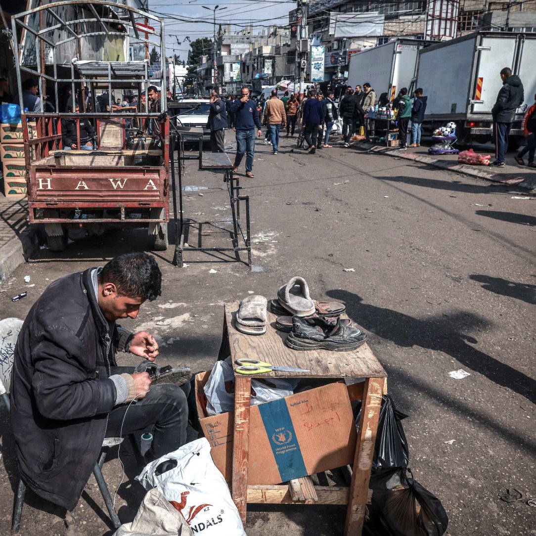 Nell'immagine c'è una strada affollata con diverse persone che si muovono. In primo piano, un uomo è seduto su una sedia accanto a un tavolo, occupato a riparare delle scarpe. Attorno a lui ci sono alcuni oggetti e sacchetti. Sullo sfondo si vedono veicoli e altre persone che camminano. L'atmosfera è vivace e caotica.
