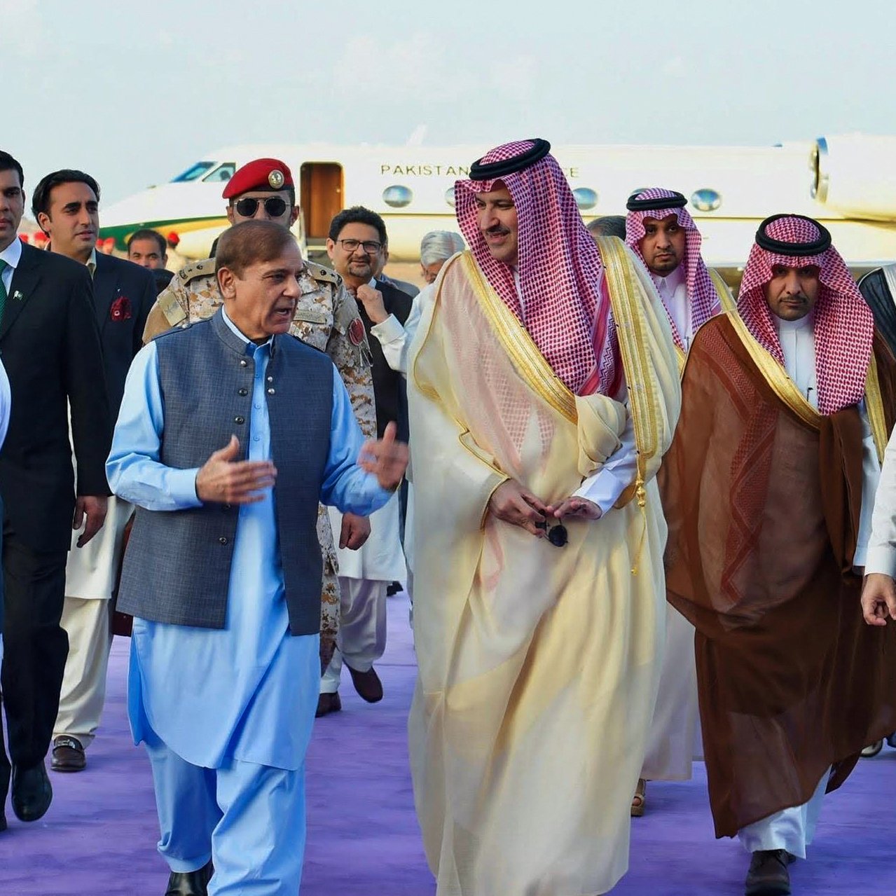 En la imagen, se puede ver a un grupo de hombres en un evento formal. Uno de ellos, vestido con una túnica azul, parece estar conversando animadamente mientras camina. Los demás, vestidos con trajes tradicionales y de gala, incluyen a personas con vestimenta árabe y otros trajes formales. Están rodeados de un ambiente elegante, con un jet privado y una alfombra morada en el fondo. La escena sugiere una ocasión diplomática o una visita de estado.