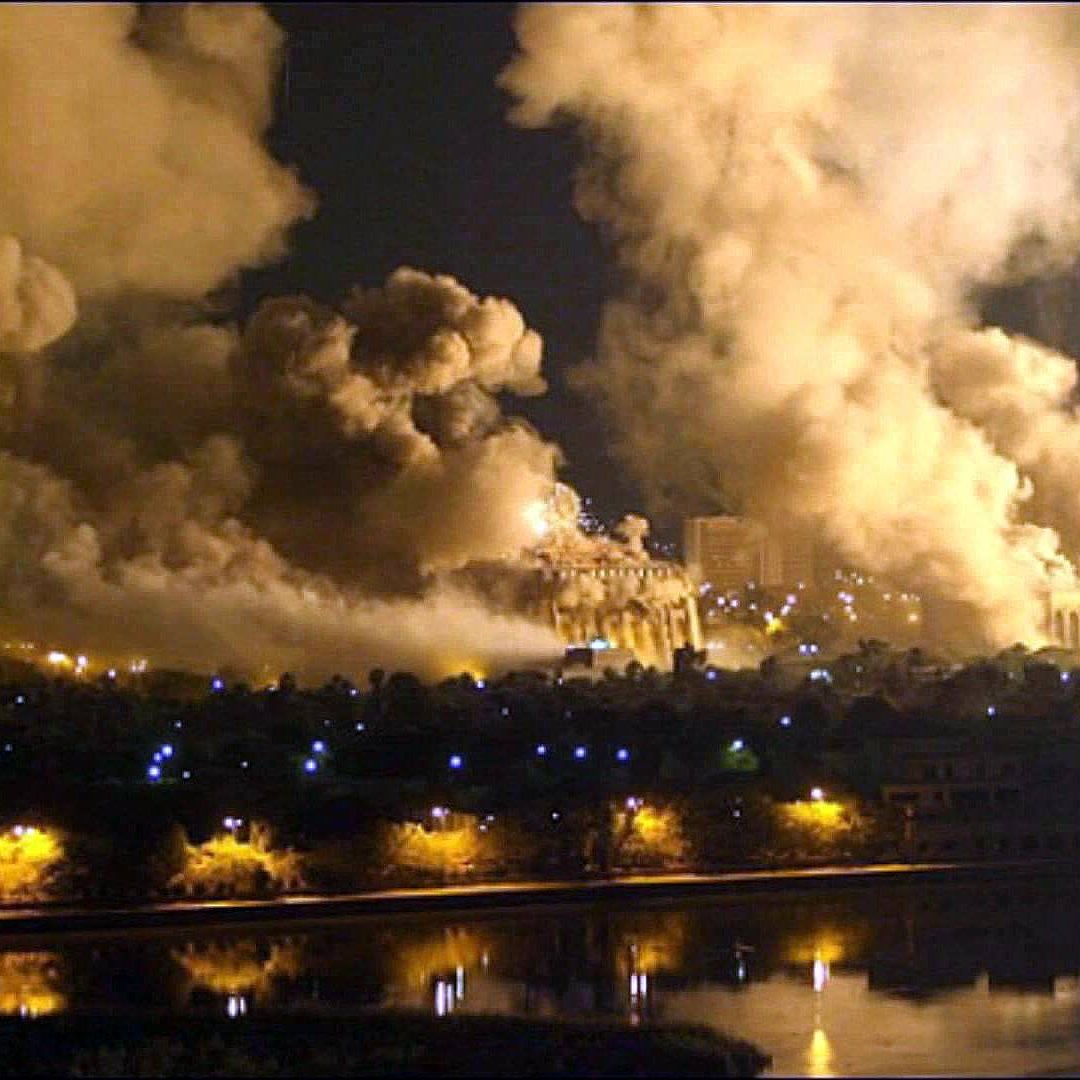 L'image montre une scène nocturne dramatique avec des nuages de fumée épais s'élevant vers le ciel. On peut voir des structures lumineuses en arrière-plan, suggérant que des bâtiments sont présents, mais ils sont partiellement obscurcis par la fumée. La réflexion des lumières sur une surface d'eau au premier plan ajoute une atmosphère sombre et intense à la composition. L'ensemble évoque une situation de conflit ou de désastre.