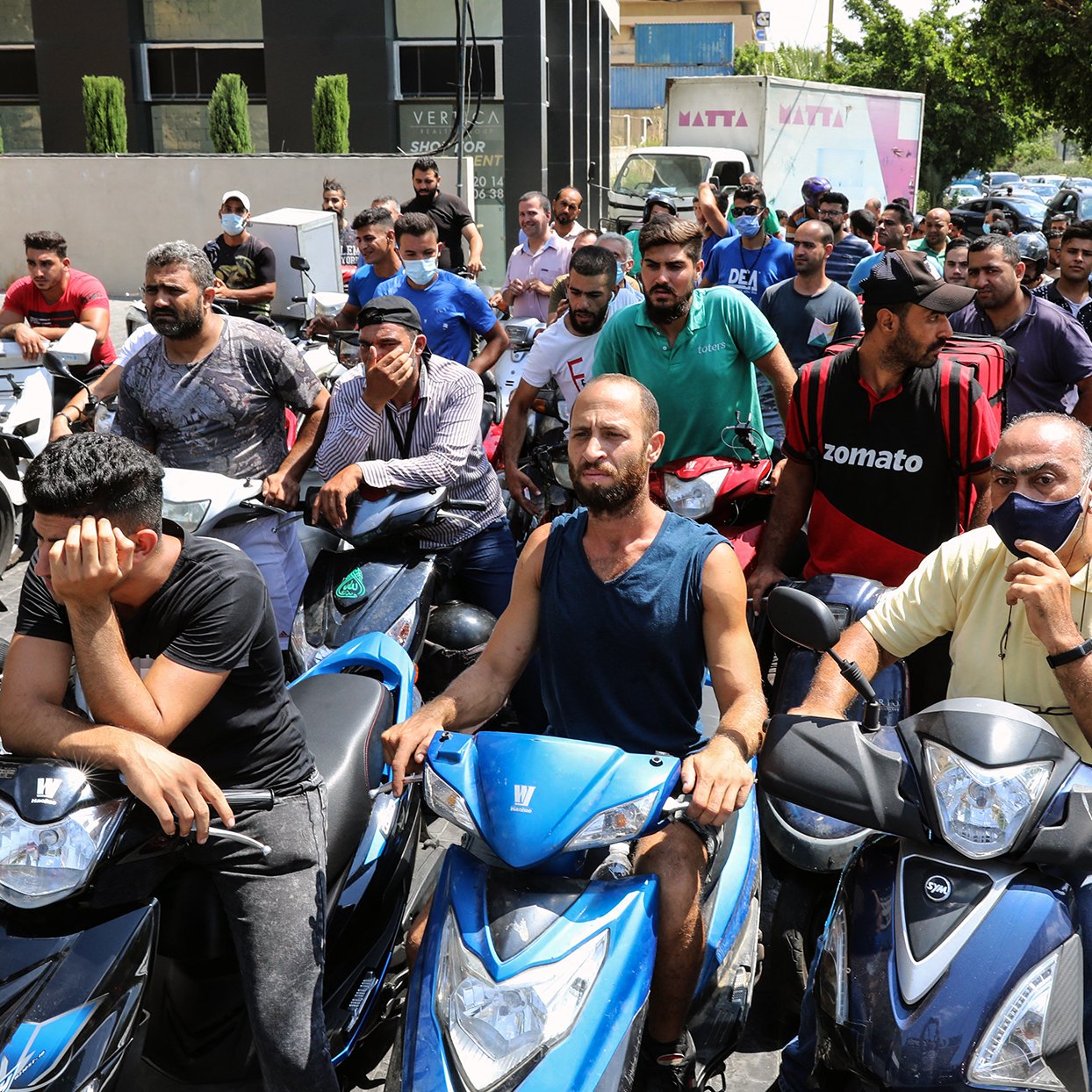 L'immagine mostra un gruppo di persone in fila, molte delle quali sono sedute su motociclette. Sembra che stiano aspettando per ricevere un servizio o un prodotto, probabilmente in un contesto urbano. Le espressioni dei volti variano: alcuni sembrano impazienti, mentre altri appaiono più rilassati. Sullo sfondo si vedono edifici e un'auto, contribuendo a creare un'atmosfera di vita cittadina.