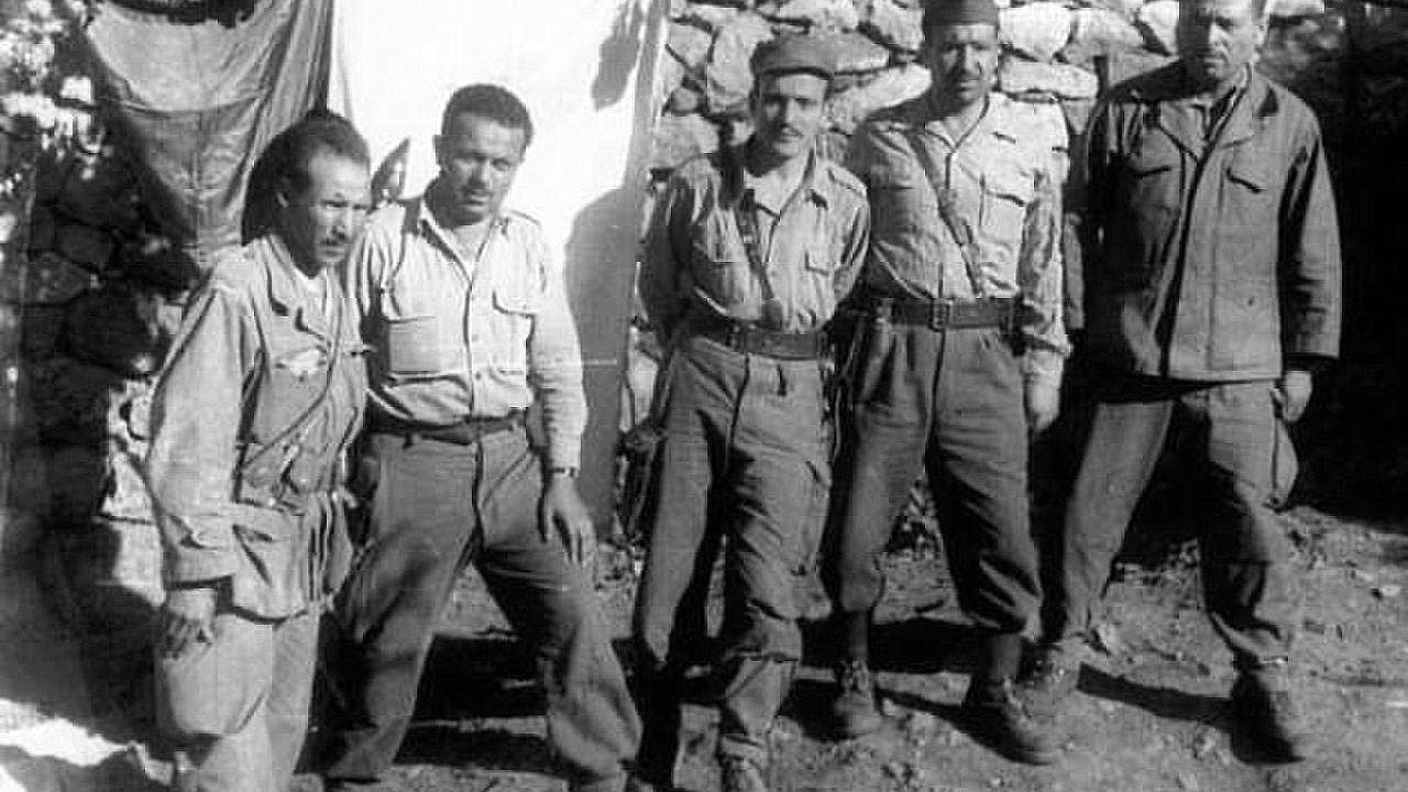 L'image montre un groupe de cinq hommes, probablement des soldats, posant ensemble devant une structure en pierre. Ils portent des uniformes militaires, composés de chemises à manches longues et de pantalons. Les expressions de leurs visages sont sérieuses et déterminées. En arrière-plan, on peut voir une toile suspendue et des pierres qui forment un mur. L'ambiance semble être celle d'un camp militaire.