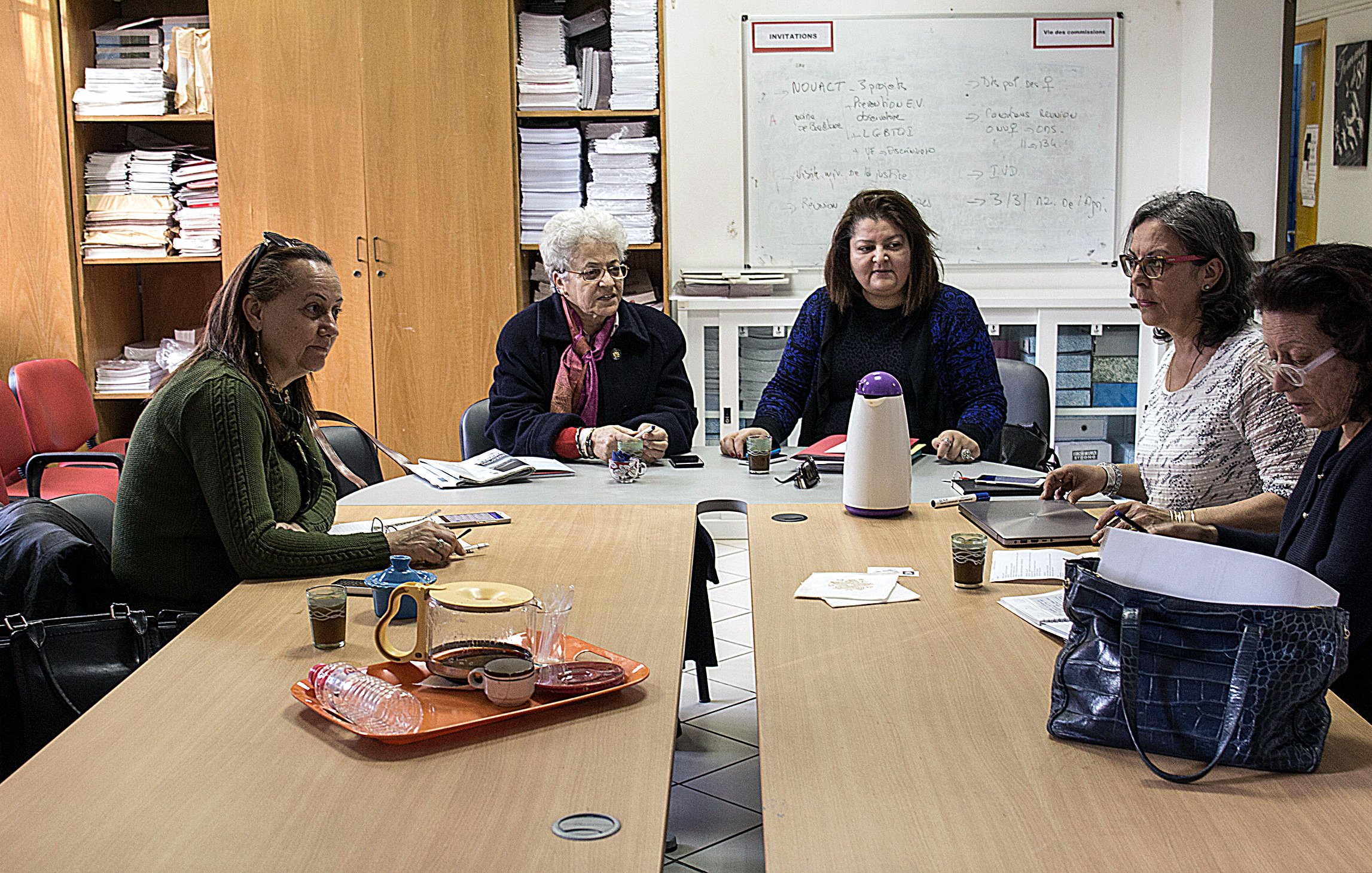 The image depicts a group of five women sitting around a table in a room filled with shelves and papers. They appear to be engaged in a discussion or work-related activity. The table has some items on it, including drinks and papers, suggesting a collaborative atmosphere. The women are looking at each other, indicating communication and interaction among them. The setting seems to be an office or a meeting room.