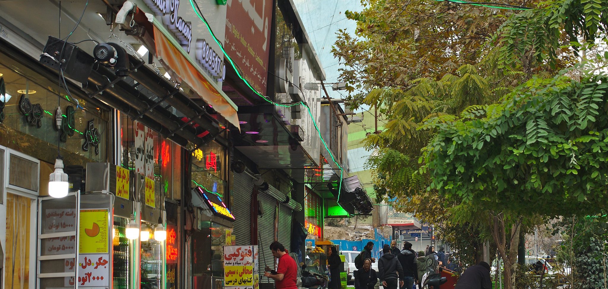 A busy street lined with shops, people walking, and colorful lights among trees.