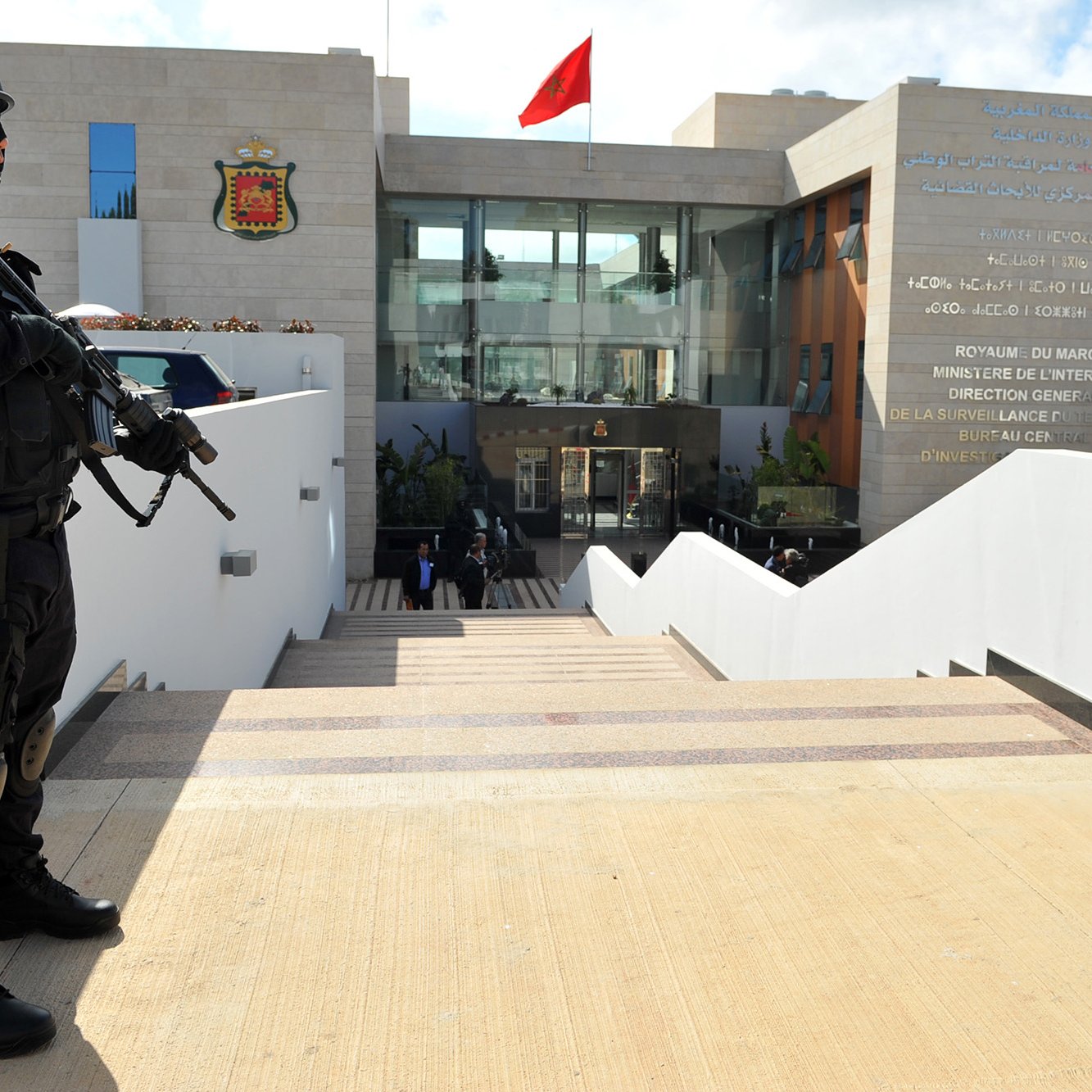The image depicts a heavily armed police officer or security personnel standing at attention outside a building. The officer is dressed in black tactical gear and is holding a firearm. In the background, there is a modern structure with a large entrance, and the Moroccan flag is prominently displayed on top. The building appears to be a government facility, likely related to security or law enforcement, as indicated by the presence of the armed guard. The setting features stairs leading up to the entrance, with a clean, contemporary design.