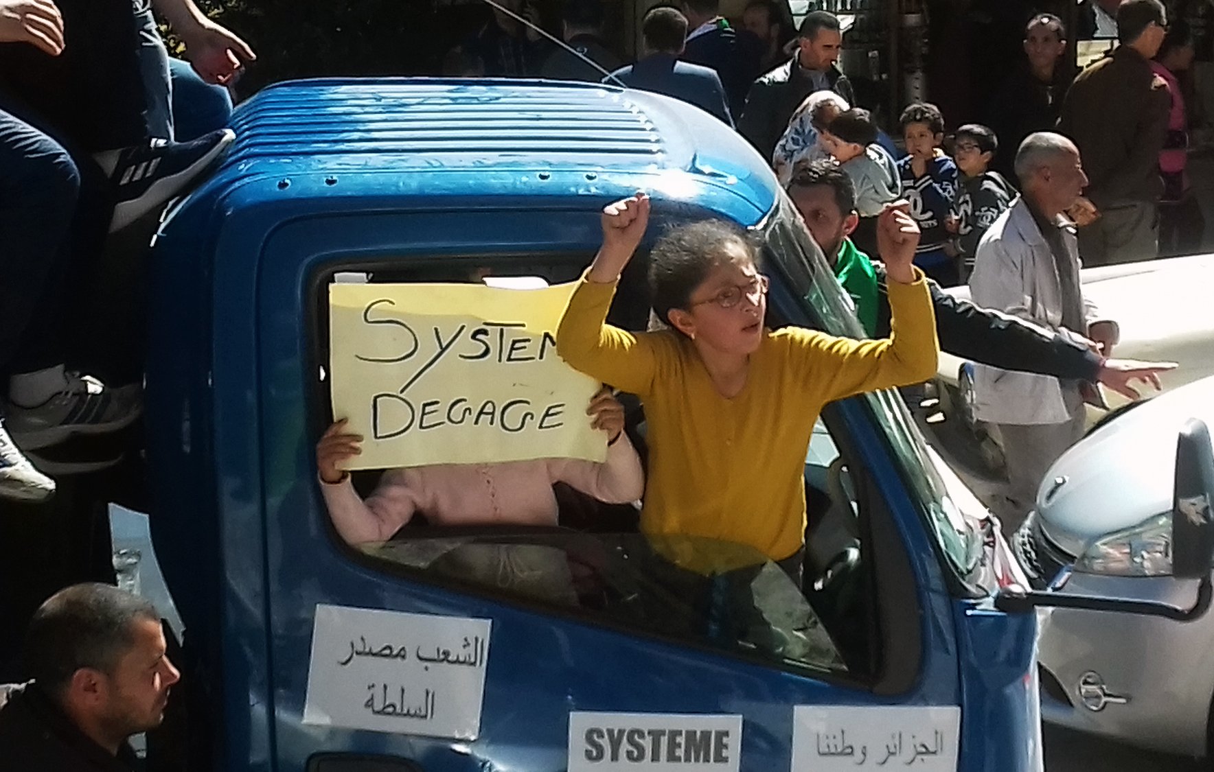L'image montre un groupe de personnes rassemblées autour d'un camion. Une femme, avec les bras levés, semble exprimer sa frustration ou son indignation. À côté d'elle, un enfant tient une pancarte avec l'inscription "Système Dégage". Le camion est décoré de quelques autres affiches, et la scène se déroule probablement lors d'une manifestation ou d'une protestation. L'atmosphère semble énergique et engagée, avec d'autres personnes en arrière-plan qui participent à la même dynamique.