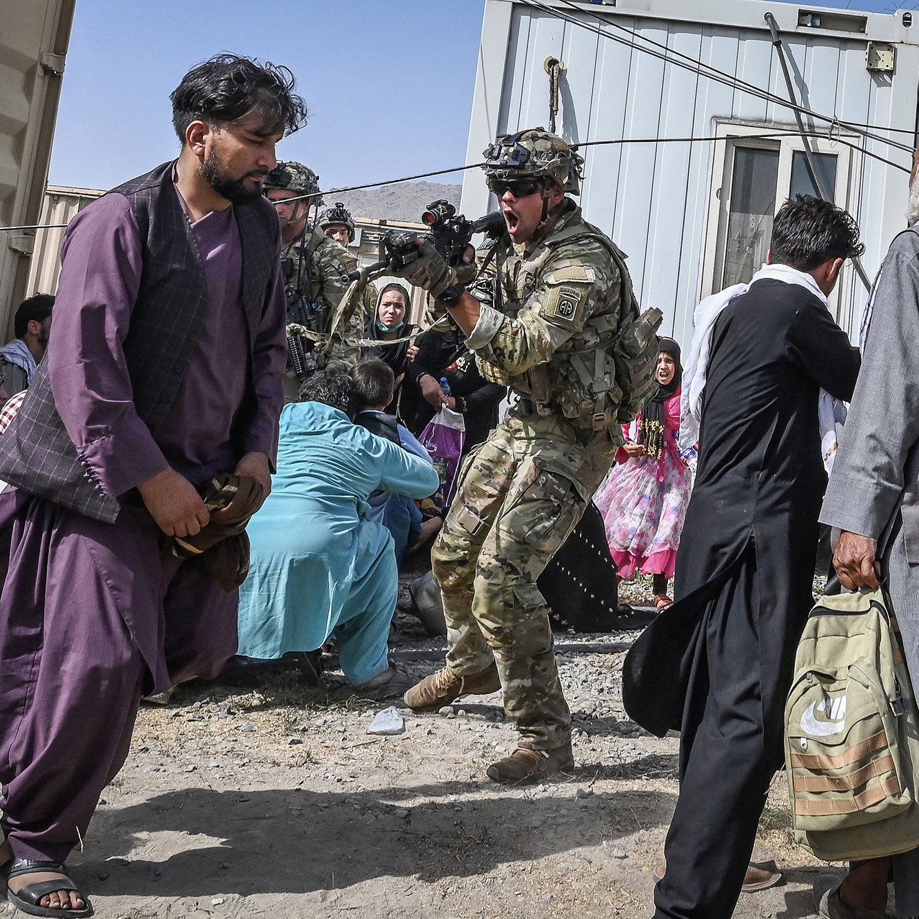 در این تصویر، یک سرباز در حالتی آماده به کار دیده می‌شود و سلاحی در دست دارد. او به نظر می‌رسد در حال محافظت از افراد در یک محیط شلوغ و اضطراب‌آور است. چندین نفر در اطراف او دیده می‌شوند، شامل مردانی با لباس‌های سنتی و زنانی که به خانه‌های محلی نزدیک هستند. فضای تصویر حاکی از یک وضعیت بحرانی و نیاز به امنیت است. مناطق اطراف نیز به نظر می‌رسد بخشی از یک کمپ یا منطقه استقرار موقت باشد.
