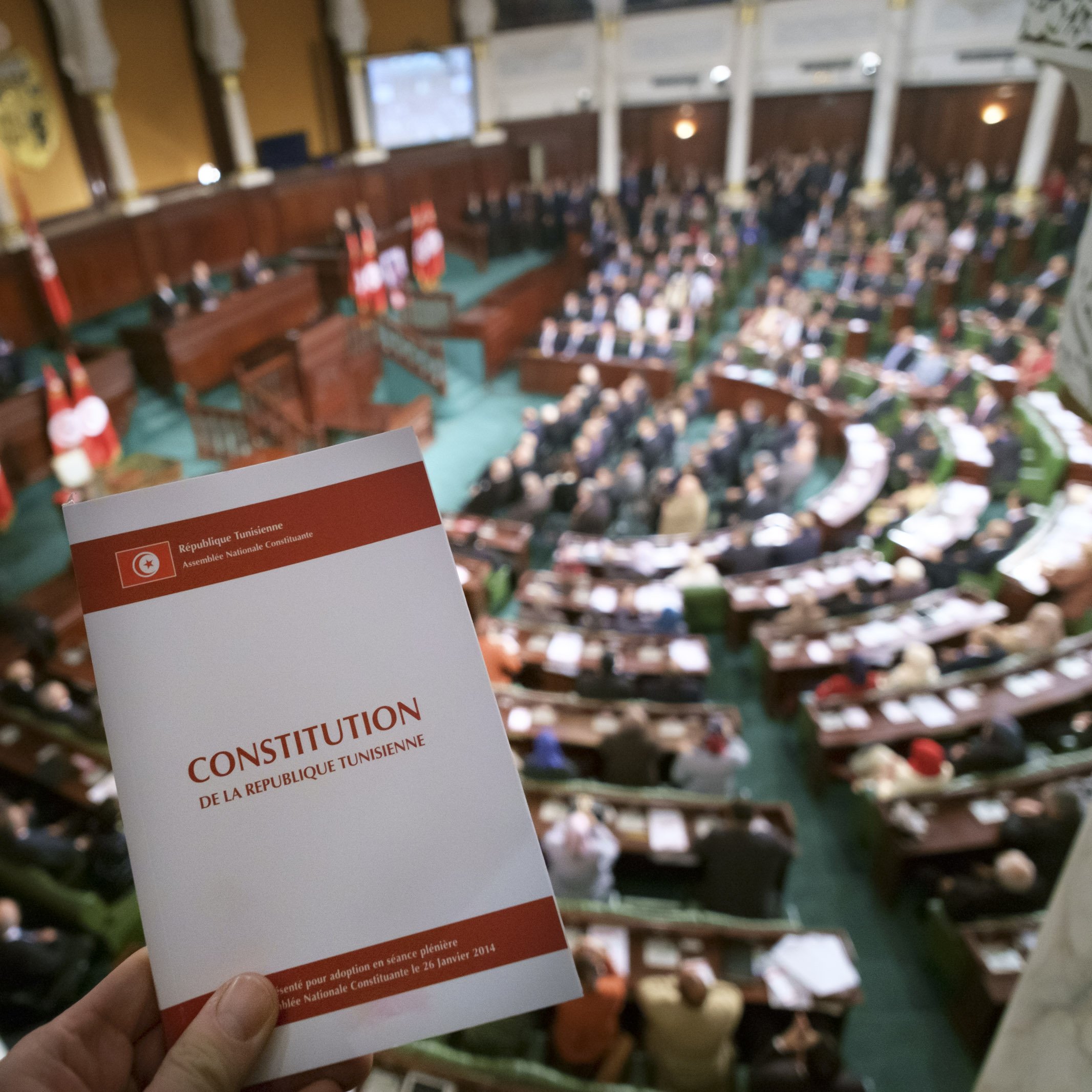 تظهر في الصورة قاعة البرلمان التونسي، حيث يوجد مجموعة كبيرة من النواب والموظفين في الجلسة. في المقدمة، هناك شخص يحمل نسخة من الدستور التونسي. الخلفية مليئة بالعلم التونسي، مما يدل على أهمية الحدث الذي يتم مناقشته في القاعة. التصميم الداخلي للقاعة يبدو أنيقًا، مع تفاصيل معمارية تقليدية.