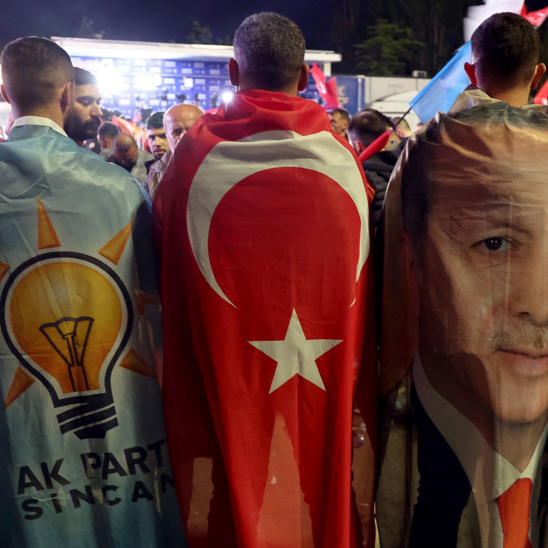 L'immagine mostra un gruppo di persone che indossano mantelli colorati. Due di loro hanno i mantelli decorati con il simbolo del partito AKP (Partito della Giustizia e dello Sviluppo) e la bandiera turca. Inoltre, uno dei mantelli presenta l'immagine di una figura pubblica, probabilmente un leader politico turco. Sullo sfondo si possono vedere altre bandiere e un'atmosfera di festa o celebrazione.