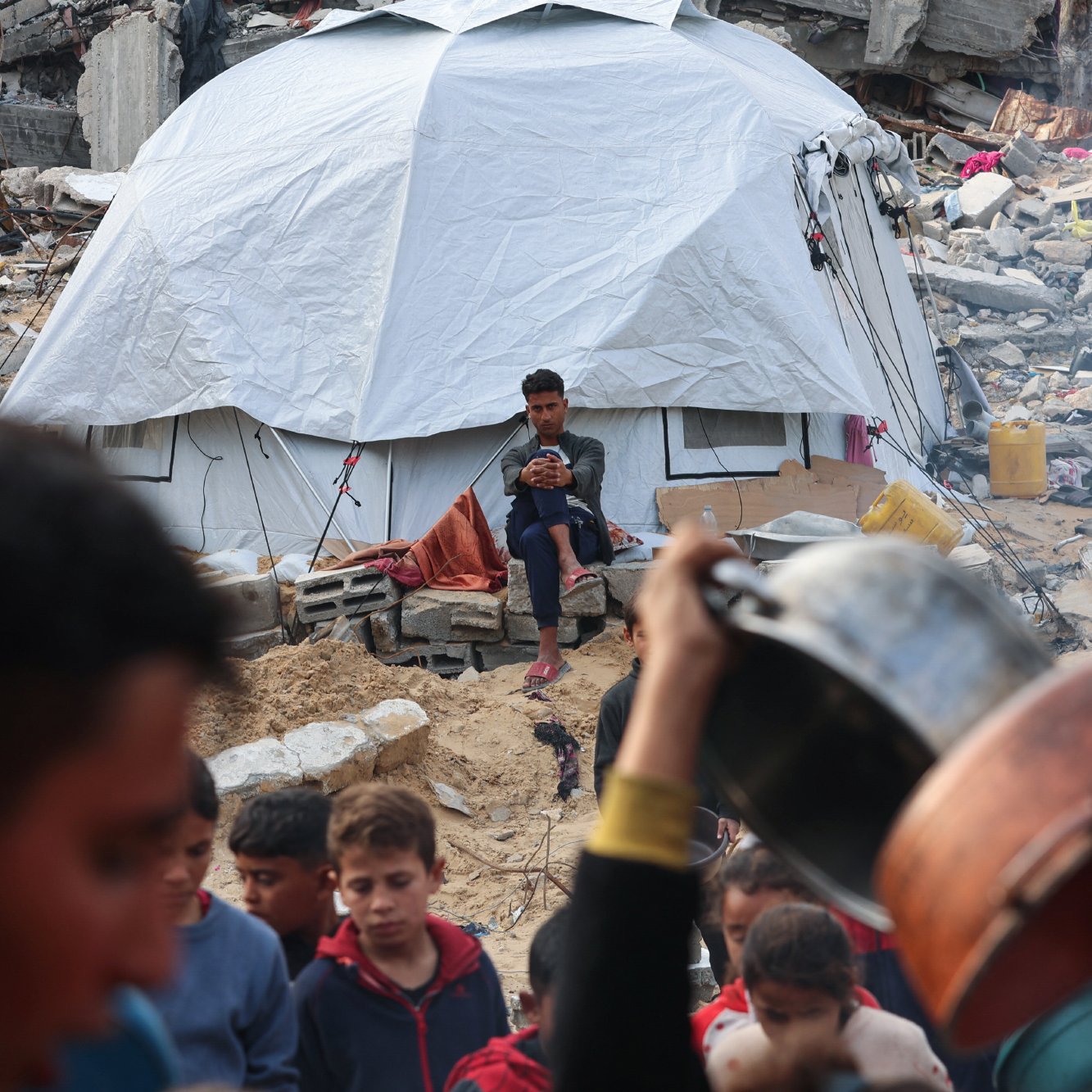 Nell'immagine si vede una tenda bianca in un contesto di distruzione, con rovine e macerie intorno. Un ragazzo è seduto davanti alla tenda, mentre in primo piano ci sono persone che si muovono e tengono stoviglie. L'atmosfera sembra di emergenza e difficoltà.