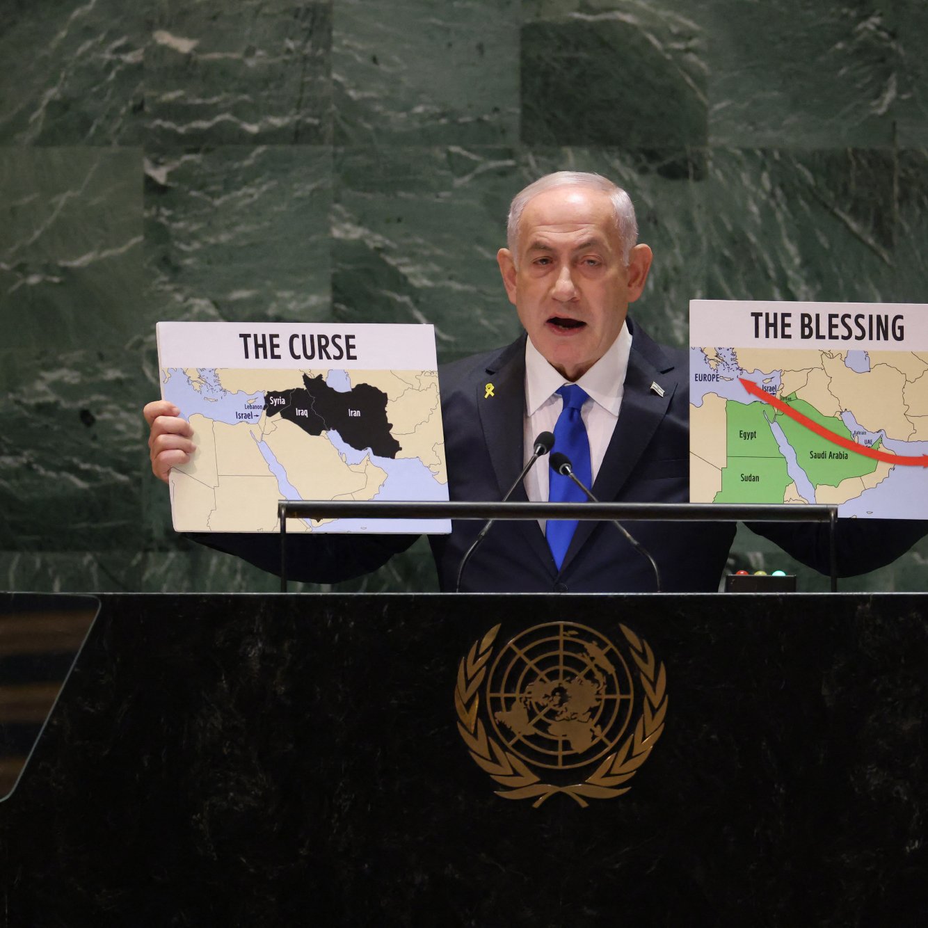 در این تصویر، یک مرد در حال صحبت در پشت یک تریبون است. او دو نقشه را در دست دارد که بر روی یکی از آن‌ها نوشته شده "نفرین" و بر روی دیگری "برکت". مرد به سمت حضار اشاره می‌کند و به نظر می‌رسد در حال بیان یک موضع سیاسی یا اجتماعی مهم است. پس‌زمینه تریبون نشان‌دهنده نماد سازمان ملل متحد است.