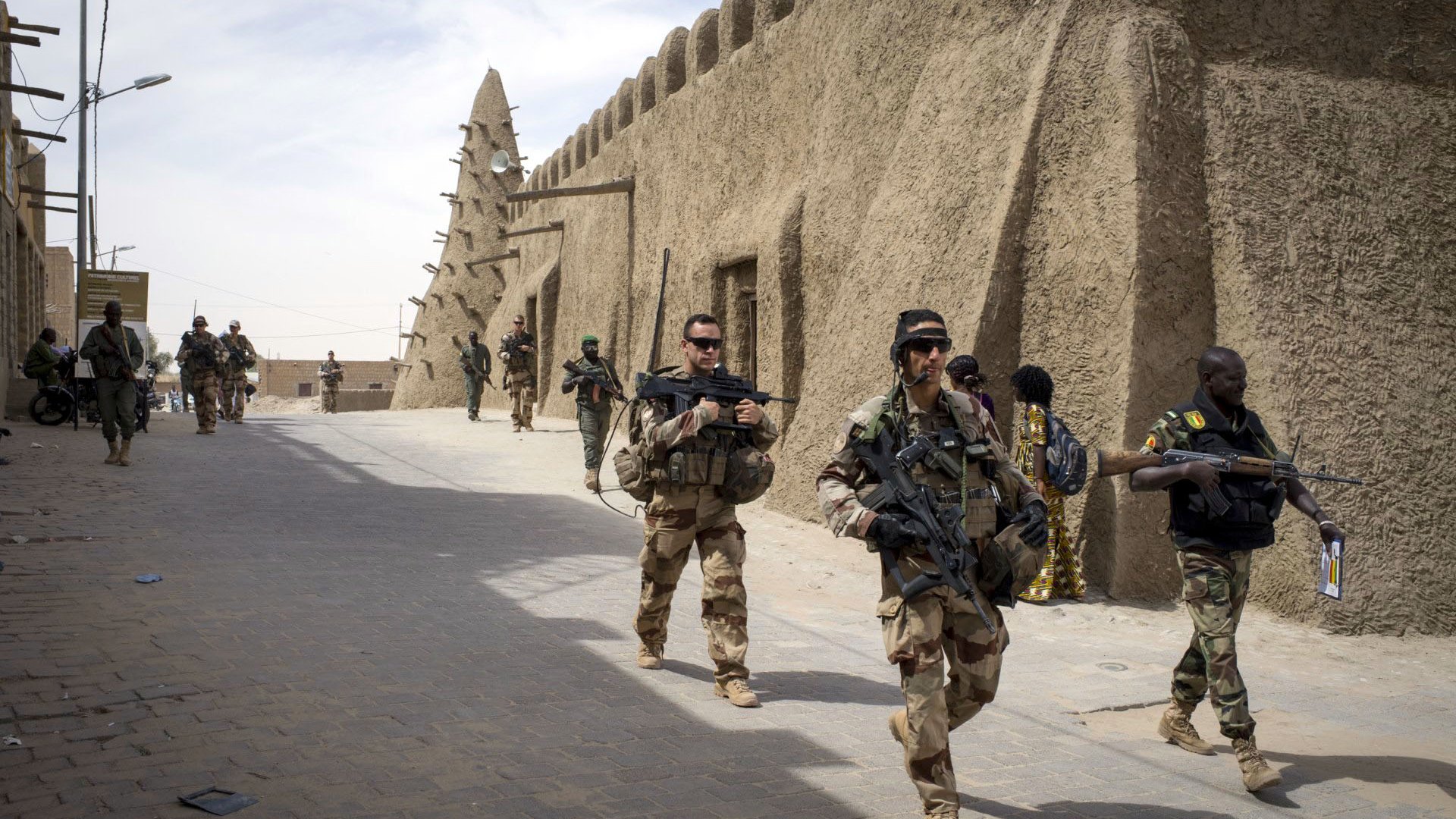 The image depicts a group of soldiers walking through a narrow street in a fortified area. They are dressed in military gear and armed with rifles. The architecture features traditional, mud-brick structures, which create a historical and cultural backdrop. The scene conveys a sense of military presence and activity, with more soldiers seen in the background, indicating a coordinated operation or patrol. The atmosphere appears to be tense and vigilant, typical of a military environment.