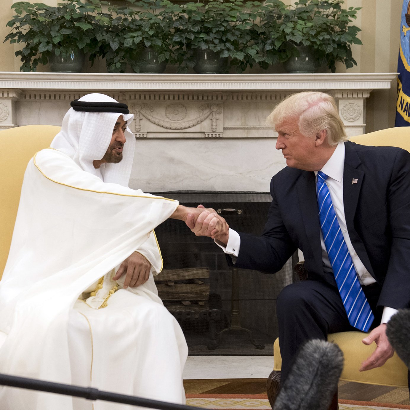 The image shows a meeting between two men, one wearing a white traditional robe and headscarf, and the other in a suit with a blue tie. They are engaged in a handshake, indicating a friendly interaction. The setting appears to be an official office, possibly the White House, with flags and decorative elements in the background. There is a focus on their handshake, showcasing a moment of diplomacy.
