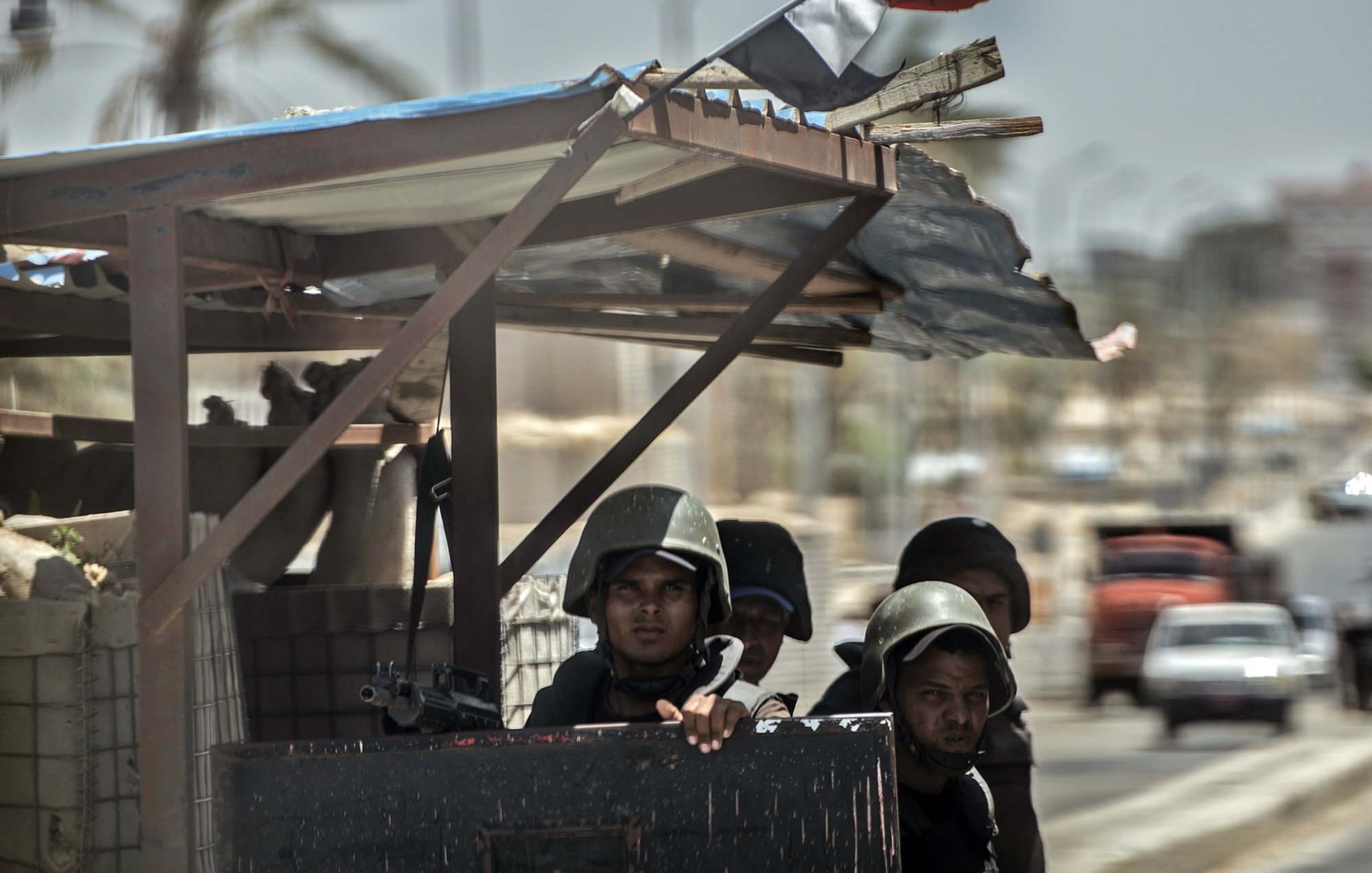 La imagen muestra a un grupo de soldados en un vehículo militar o en un puesto de control. Están vestidos con uniformes de combate y cascos, y algunos de ellos parecen estar armados. Al fondo, se pueden ver árboles y edificaciones, así como un cielo despejado. También hay un pequeño trozo de una bandera ondeando. La escena sugiere un ambiente de vigilancia o seguridad en una zona urbana.