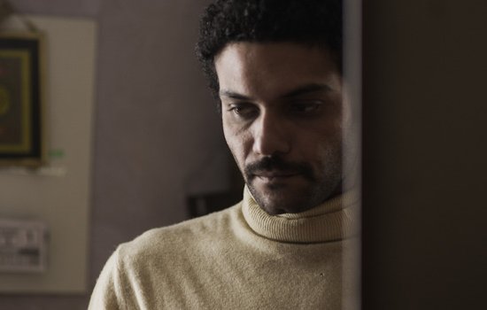 L'image montre un homme avec des cheveux bouclés et une moustache, portant un pull à col roulé de couleur claire. Il semble pensif, regardant vers le bas, avec une expression de réflexion ou de sérieux. En arrière-plan, on aperçoit des éléments de décoration intérieure, mais les détails sont flous. L'atmosphère de l'image dégage une certaine intimité ou mélancolie.