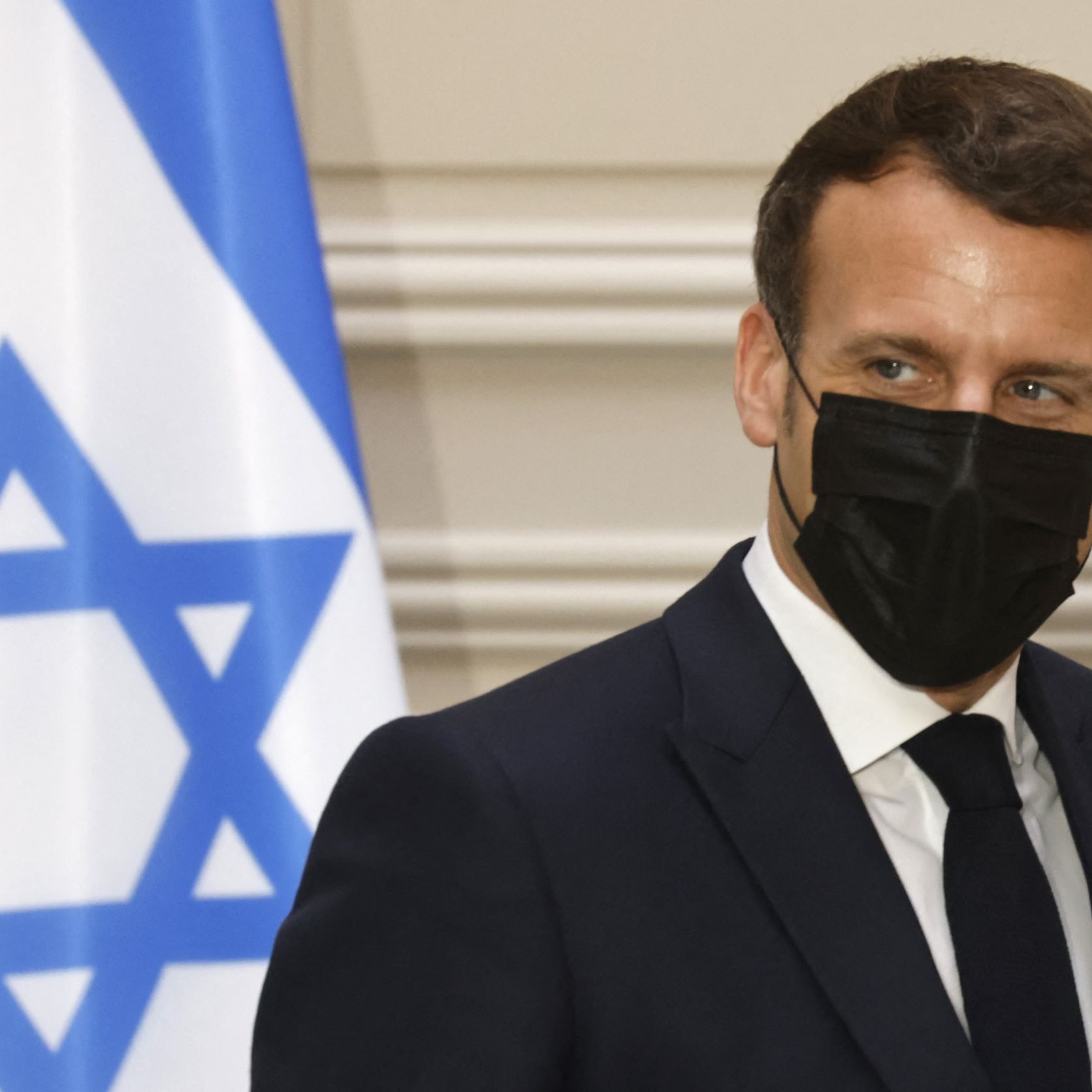 En la imagen se puede ver a un hombre con un traje oscuro y corbata, que lleva una mascarilla negra. Está de perfil y parece estar en un evento oficial. Al fondo hay una bandera con la estrella de David, indicando que el contexto puede estar relacionado con Israel. La expresión del hombre es seria y concentrada.