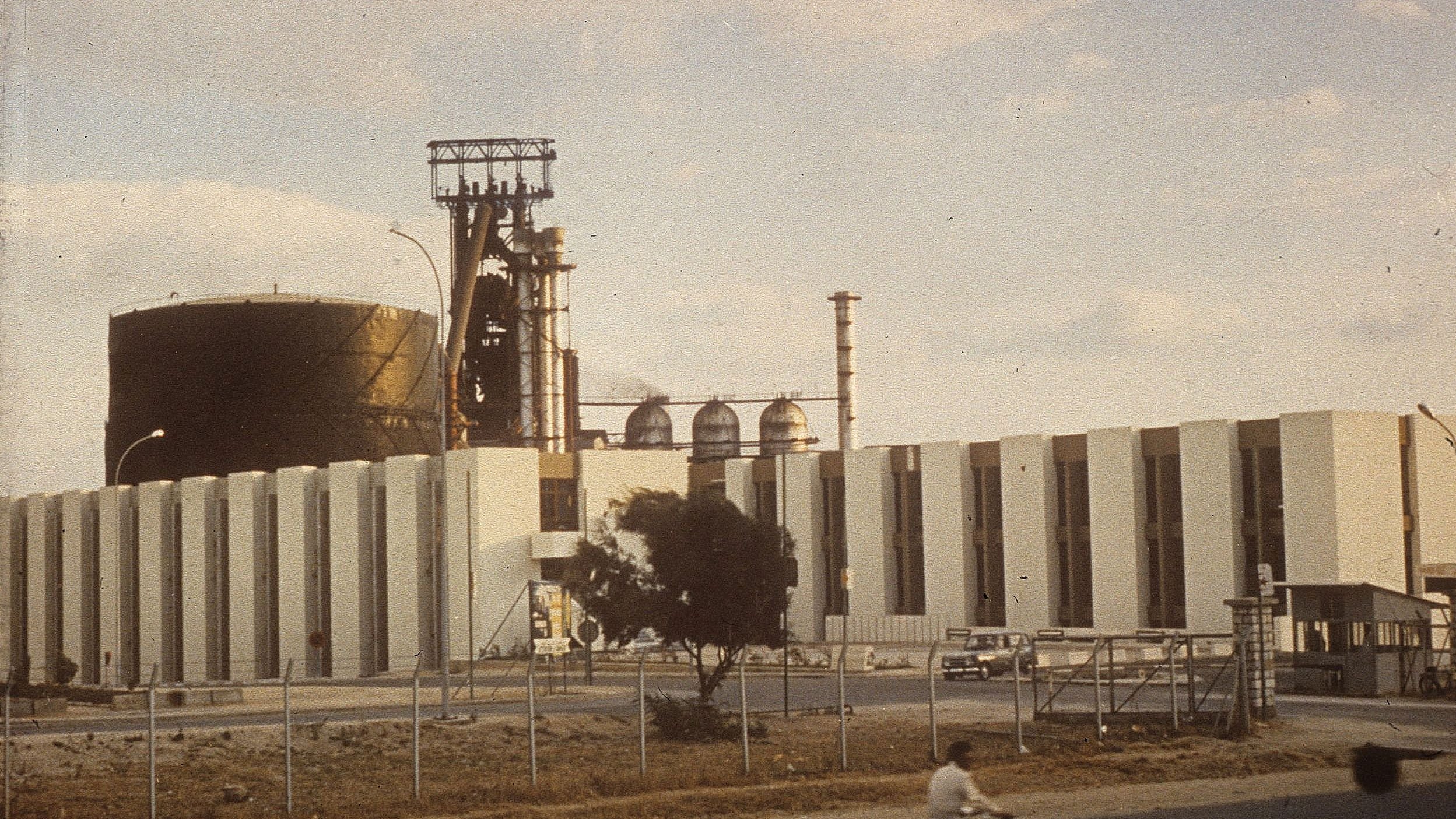 L'image montre une grande installation industrielle. On peut voir un grand réservoir noir et une structure avec des cheminées et des tubes. La façade de l'immeuble est faite de blocs blancs. À l'avant, il y a une route, et on aperçoit une personne à vélo sur celle-ci. L'environnement semble être un site industriel, avec quelques arbres et une clôture qui délimite la zone. Le ciel est partiellement nuageux, ce qui donne une ambiance légèrement vintage à la scène.