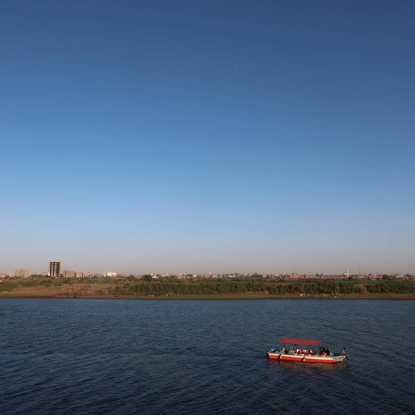 L'image montre une scène paisible d'un cours d'eau avec un ciel dégagé et sans nuages. Au centre, on peut voir un petit bateau naviguant sur les eaux calmes. À l'arrière-plan, on distingue une ligne d'horizon urbaine, avec des bâtiments et des arbres le long de la rive. L'ambiance générale est tranquille et ensoleillée, suggérant une belle journée.