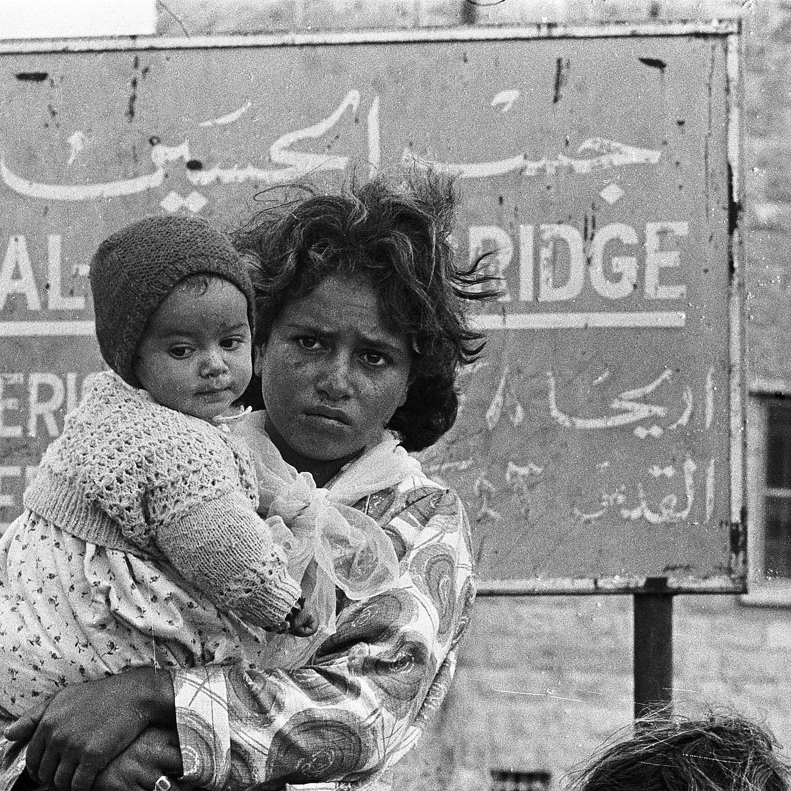 این تصویر یک کودک را نشان می‌دهد که در آغوش یک زن ایستاده است. زن ظاهراً نگران و جدی به نظر می‌رسد و کودک در آغوشش پایش را به دور او حلقه کرده است. پس‌زمینه تصویر شامل یک تابلو با نوشته‌ها به زبان عربی و انگلیسی است که نام مکانی را نشان می‌دهد. حالت چهره و لباس‌های آن‌ها نشان‌دهنده سختی‌های احتمالی در زندگی است. تصویر به وضوح وضعیت انسانی و احساسات عمیق را منتقل می‌کند.