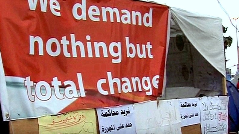L'image montre une grande pancarte rouge avec le texte "We demand nothing but total change" en anglais, ce qui signifie "Nous exigeons rien d'autre qu'un changement total". En dessous de cette pancarte, on peut voir plusieurs autres affiches, probablement en arabe, qui expriment également des demandes ou des revendications. L'ensemble de la scène évoque un lieu de manifestation ou de protestation, suggérant des aspirations à un changement significatif.