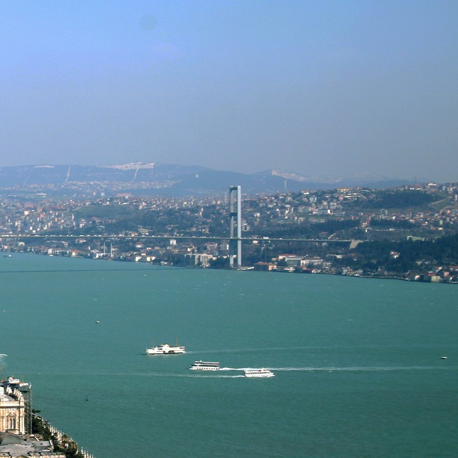 تصویر نمای کلی از یک شهر در کنار یک دریاچه یا رودخانه را نمایش می‌دهد. در سمت چپ تصویر، ساختارها و ساختمان‌های مختلفی دیده می‌شود که نشان‌دهنده مناطق مسکونی و تجاری است. در پس‌زمینه، پل بزرگی وجود دارد که دو طرف آب را به هم متصل می‌کند. آسمان آبی و صاف است و دورنمای تپه‌ها و شهر در پس‌زمینه به وضوح قابل مشاهده است. همچنین، برخی قایق‌ها در سطح آب در حال حرکت هستند. این تصویر حس سرزندگی و زیبایی طبیعی منطقه را منتقل می‌کند.