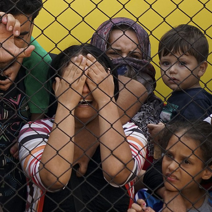 L'image montre un groupe de personnes derrière une clôture en métal. Au centre, une femme semble très émue et cache son visage avec ses mains, tandis que plusieurs enfants et adultes l'observent avec des expressions d'inquiétude et de tristesse. Le contraste entre la clôture et les visages des personnes souligne un sentiment d'angoisse et de vulnérabilité. Les couleurs de l'arrière-plan sont vives, mais l'atmosphère ressentie est lourde et chargée d'émotions.