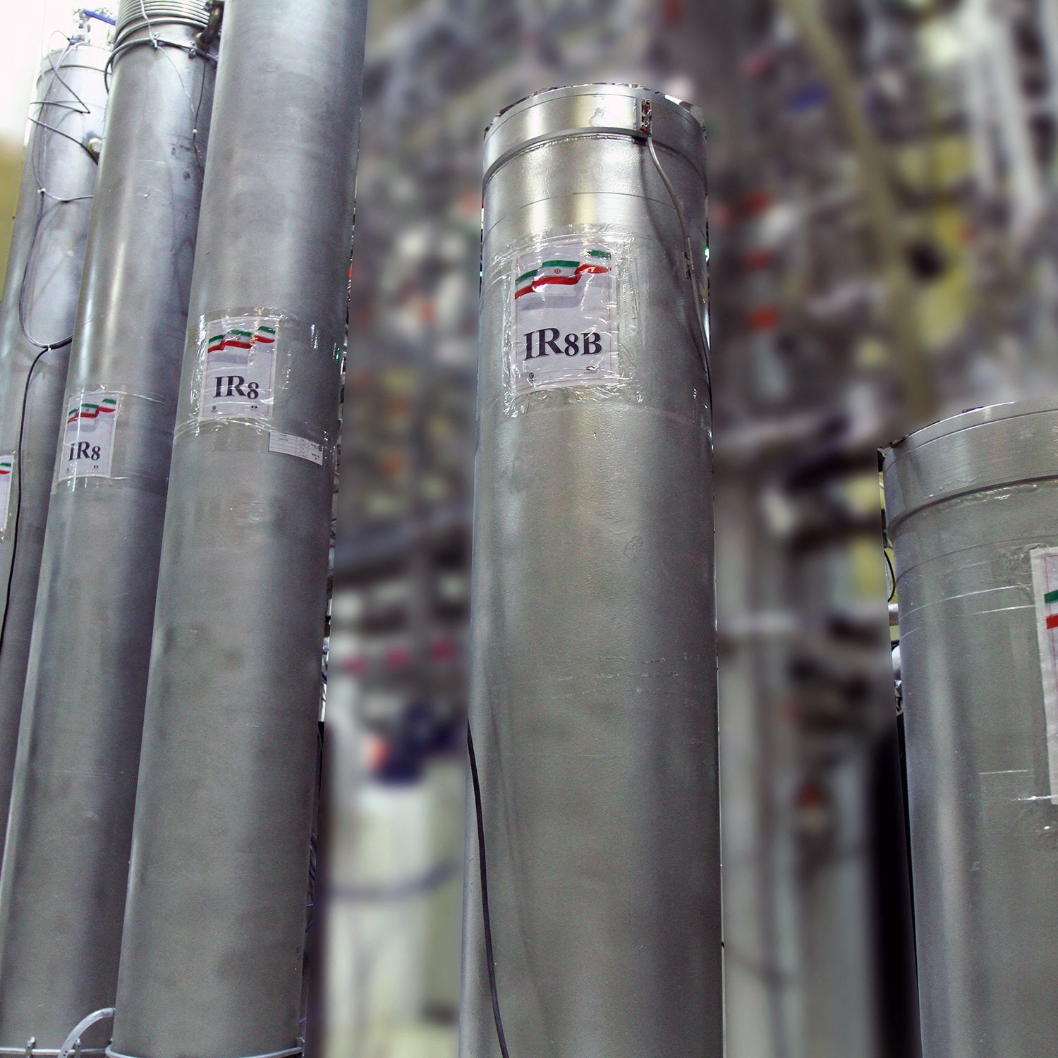 La imagen muestra varios cilindros metálicos dispuestos en un laboratorio o instalación industrial. Cada cilindro tiene una etiqueta que indica "IR8" o "IR8b", probablemente refiriéndose a modelos de centrifugas. En las etiquetas se puede ver una representación de la bandera de Irán. El fondo está desenfocado, pero se pueden observar más estructuras relacionadas con el equipo técnico, sugiriendo un ambiente relacionado con la investigación o la producción.