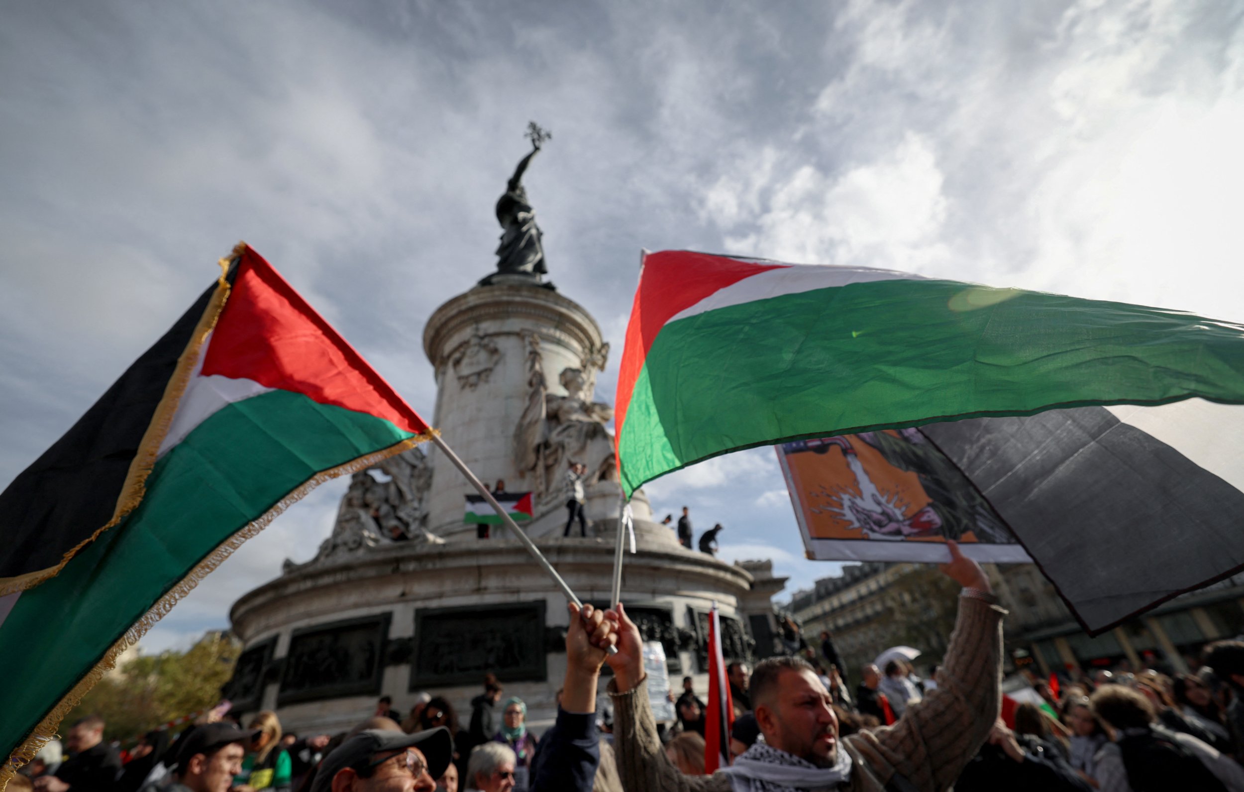 تظهر الصورة مجموعة من الأشخاص وهم يحملون أعلام فلسطينية في تجمع جماهيري. الخلفية تُظهر نصبًا تذكاريًا عظيمًا، مع سماء ملبدة بالغيوم. يبدو أن الناس متحمسون ويشاركون في فعالية ذات طابع وطني أو سياسي. الأعلام تحمل ألوان العلم الفلسطيني، مما يشير إلى دعمهم للقضية الفلسطينية.