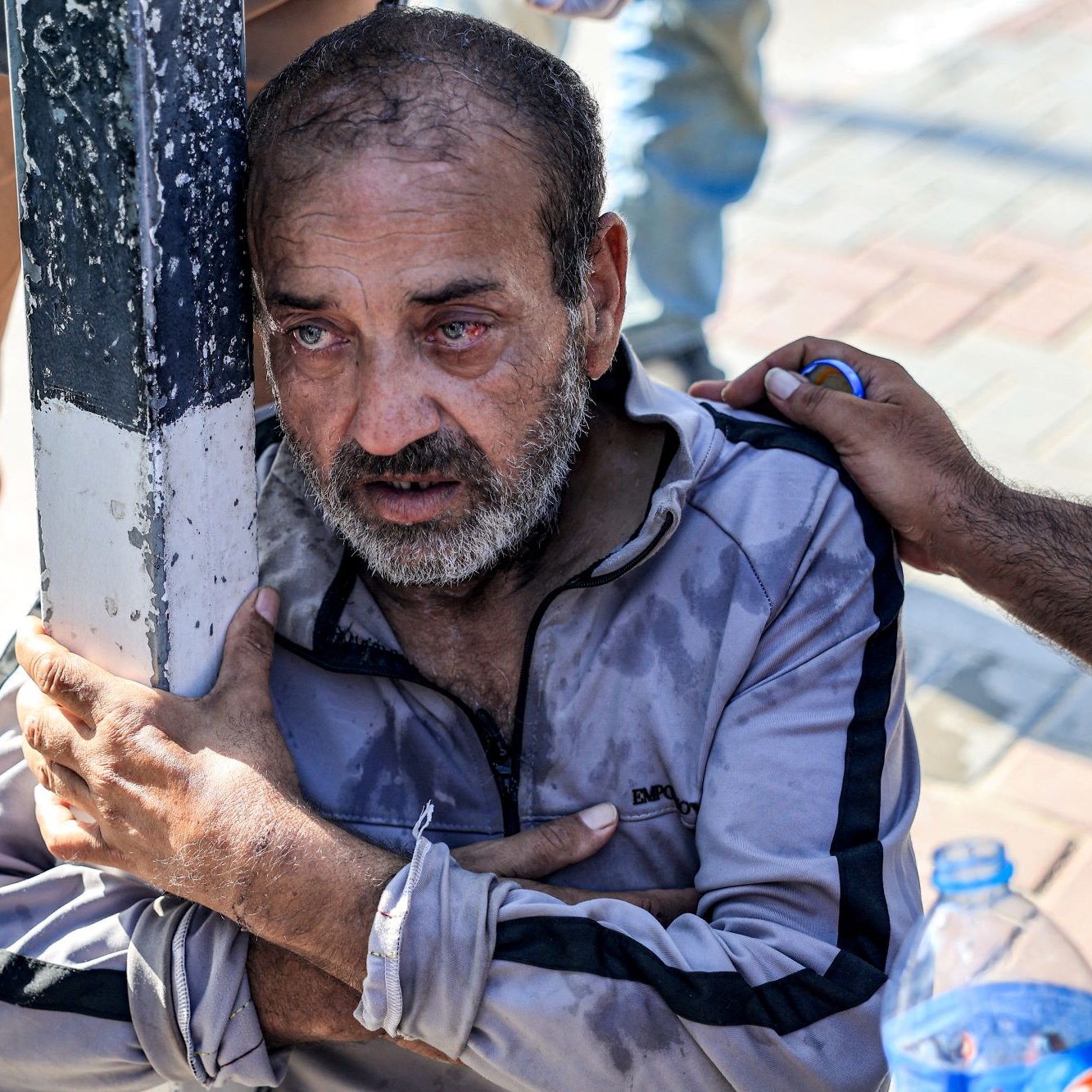 The image depicts a distressed elderly man sitting on the ground, clutching a pole for support. He appears visibly upset, with tears in his eyes and a look of despair on his face. His clothing is worn, and he seems to be in a tough situation. There are hands reaching out to him, suggesting someone is offering help or comfort. The background shows a pavement, indicating an outdoor setting. The overall atmosphere conveys a sense of vulnerability and struggle.