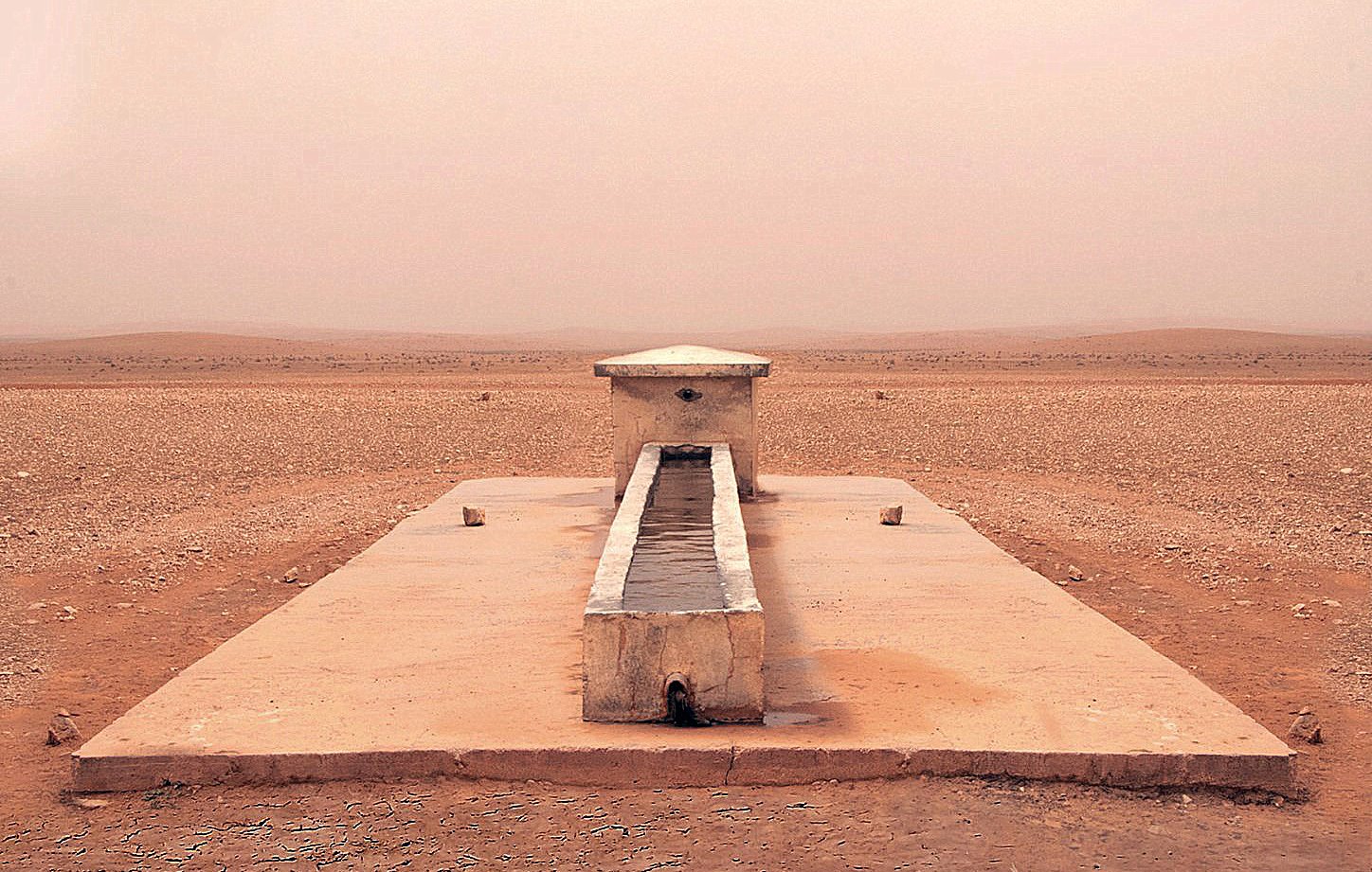 L'image montre une structure isolée située dans un paysage désertique. Au centre, il y a un long bassin en pierre, entouré par une surface plane et aride. Le sol est de couleur beige et semble sec et craquelé, tandis que l'atmosphère est légèrement brumeuse, ce qui donne une impression de chaleur et d'immensité désertique. Au loin, on aperçoit des collines ou des dunes qui se fondent avec le ciel dans une teinte douce et uniforme. L'endroit dégage une sensation de tranquillité et de solitude.