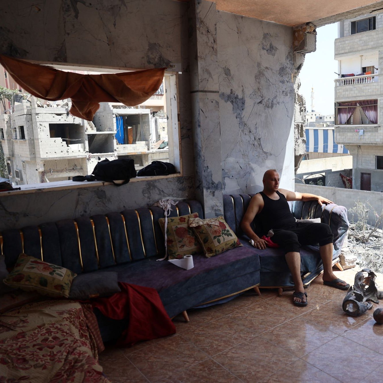 L'immagine mostra un uomo seduto su un divano in un ambiente che appare distrutto. Le pareti sono danneggiate e si possono vedere segni evidenti di distruzione. Nella stanza ci sono alcuni oggetti sparsi, come pezzi di ceramica e materiali vari. Alle spalle dell'uomo, le finestre danno su un paesaggio urbano suggerente di devastazione, con edifici danneggiati e in rovina visibili all'esterno. L'atmosfera è triste e riflette una situazione di difficoltà e lotta.