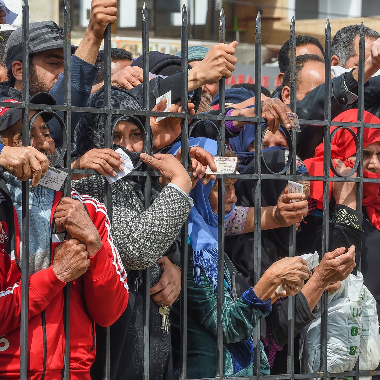 تظهر في الصورة مجموعة من الأشخاص المتجمعين خلف سياج حديدي. يرفع البعض منهم أوراقًا أو بطاقات، بينما يبدو التركيز على حركتهم وتصرفاتهم، مما يوحي بأنهم قد يكونون في انتظار شيء ما أو يحاولون الوصول إلى جهة معينة. الأجواء تعكس حالة من الترقب أو الأمل، مع تنوع في الملابس، مما يشير إلى خلفيات مختلفة.