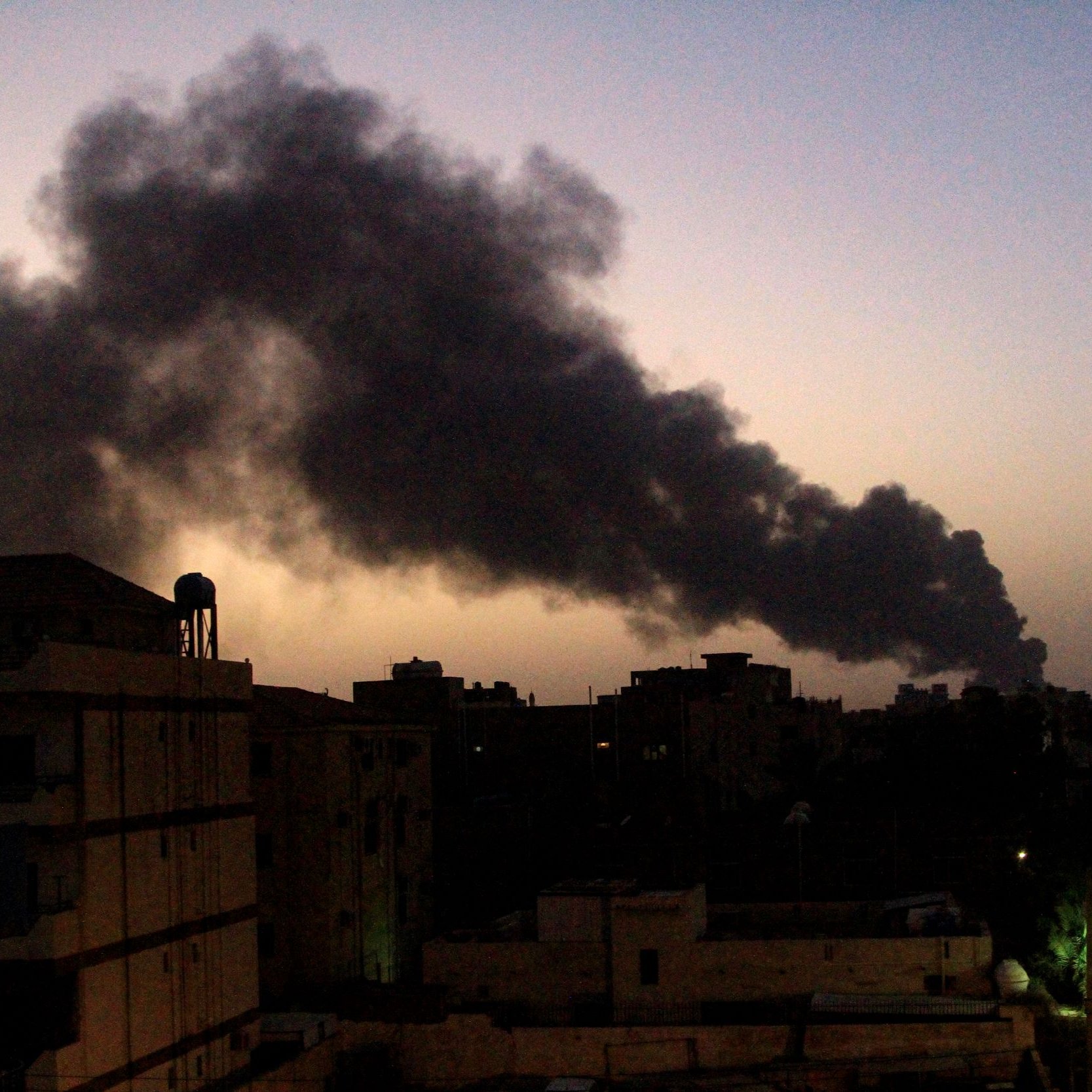 The image depicts a dusk or evening scene in an urban environment, with several buildings visible in the foreground. In the background, a substantial plume of black smoke rises into the sky, creating a stark contrast against the fading light of sunset. The smoke suggests a fire or some kind of industrial activity. The overall mood of the image conveys a sense of urgency or concern, given the presence of the smoke.