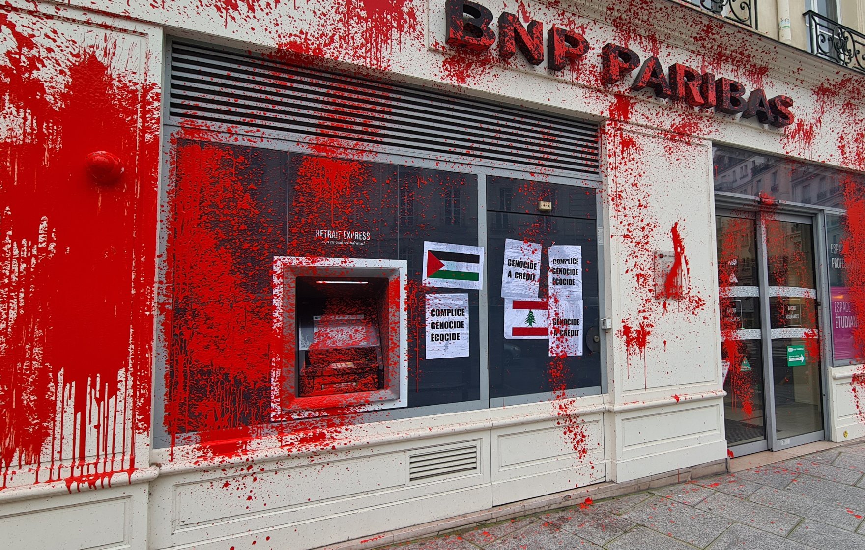 L'image montre une agence de la BNP Paribas, dont la façade est recouverte de peinture rouge. Des affiches sont visibles sur la vitrine, et l'automate de distribution de billets est également marqué par cette peinture. L'ensemble donne une impression de vandalisme ou de protestation, soulignant peut-être un message politique ou social.