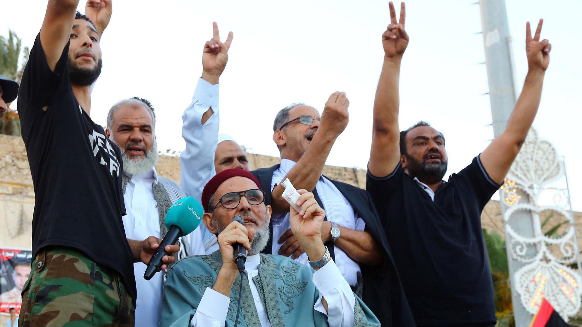 L'image montre un groupe d'hommes réunis lors d'un événement. Un homme au premier plan, portant une tenue traditionnelle, s'exprime tout en levant un symbole de paix avec ses mains. Les autres hommes en arrière-plan lèvent également les mains, probablement en signe de soutien ou de solidarité. L'ambiance semble festive et engagée, suggérant un moment de rassemblement ou de protestation. Des drapeaux ou des affiches pourraient être visibles, accentuant le contexte de l'événement.