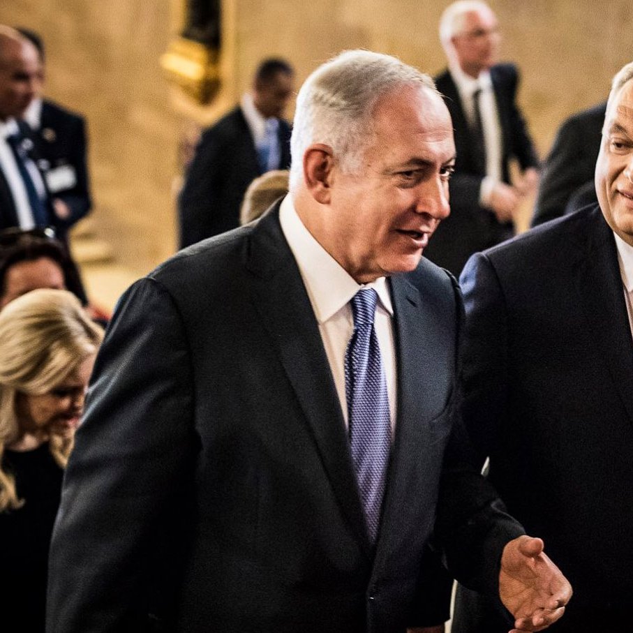 در این تصویر، دو مرد در حال صحبت و قدم زدن به سمت جلو هستند. یکی از آنها لباس سیاه و کراوات آبی به تن دارد و دیگری لباس رسمی با کراوات صورتی بر تن دارد. تعدادی از افراد دیگر نیز در پس‌زمینه وجود دارند که به نظر می‌رسد در یک مکان رسمی یا مراسم حضور دارند. فضای تصویر نشان‌دهنده یک موقعیت مهم یا رسمی است.