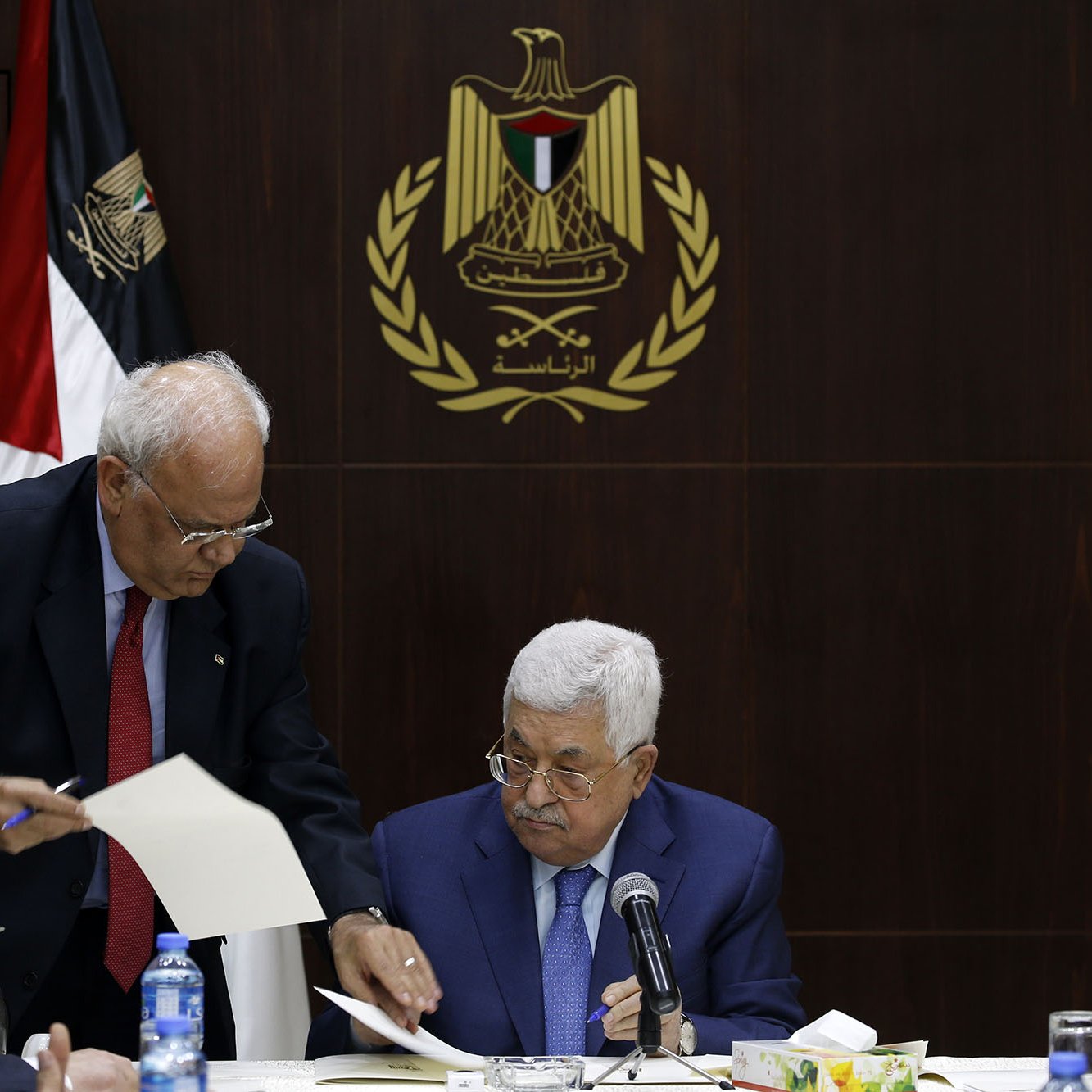 L'image montre une réunion, probablement politique, avec plusieurs hommes assis à une table. Au premier plan, un homme, probablement un leader ou un officiel, est assis regardant des documents. Un autre homme, debout à ses côtés, semble lui présenter des papiers ou des informations. En arrière-plan, on peut voir un drapeau, ainsi qu'un emblème qui semble représenter une organisation ou un gouvernement. L'ambiance générale de la scène est sérieuse et attentive, suggérant une discussion importante.