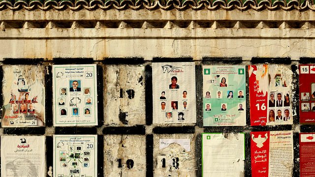 L'image montre un mur sur lequel sont affichés plusieurs documents et affiches. Ces derniers semblent être en lien avec des élections, avec des plaques présentant des photos et des noms de candidats. Certaines affiches sont abîmées et décolorées, révélant un certain vieillissement et une usure du temps. Le mur lui-même présente une texture rugueuse et des motifs décoratifs en haut. Les divers éléments placardés créent une composition visuelle chargée et informative.