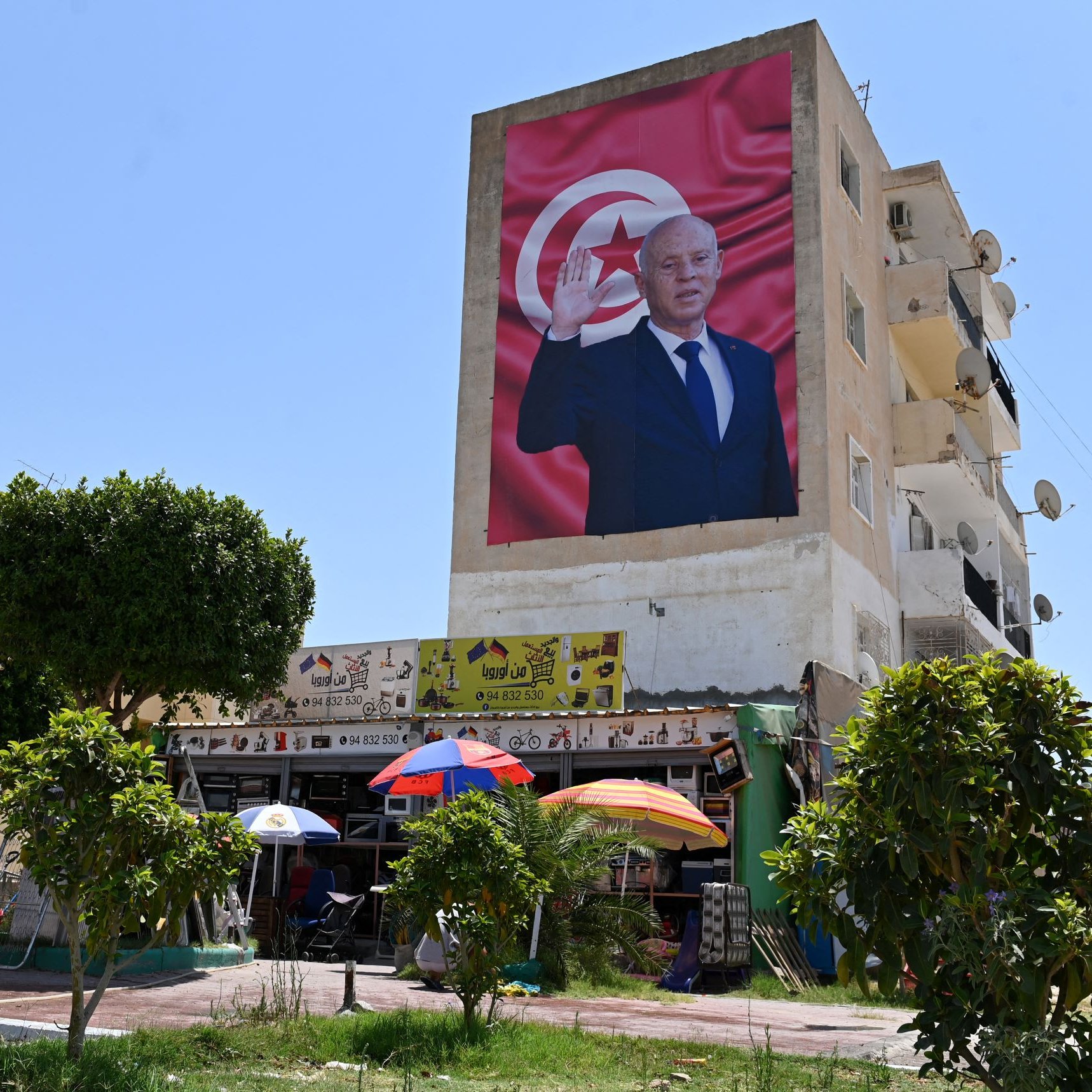 L'image montre un bâtiment avec une grande fresque représentant un homme, probablement un dirigeant, qui fait un geste de salutation. En arrière-plan, on peut voir un drapeau tunisien. Au premier plan, il y a des parasols colorés et des arbres, ainsi qu'une scène de vie qui pourrait inclure des personnes assises ou se déplaçant. Le ciel est clair et ensoleillé, ce qui donne une atmosphère lumineuse à la scène.