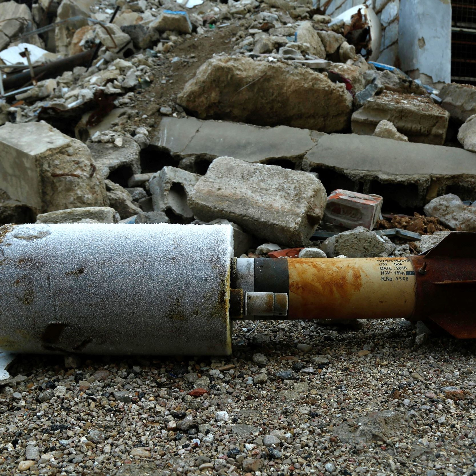 الصورة تظهر قنبلة أو جسم متفجر ملقى على الأرض، محاط بأحجار وأنقاض تشير إلى تدمير في المنطقة. يبدو أن الساحة غير مرتبة، والأرض مليئة بالردم والحطام، مما يشتت الانتباه عن الجسم المتفجر. يظهر أن هذا المكان شهد حدثًا مروعًا أدى إلى تدمير كبير.