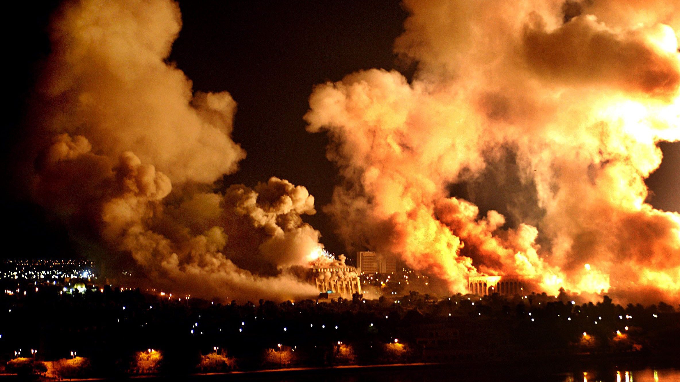 L'image montre une scène nocturne marquée par d'épais nuages de fumée et des flammes. On peut apercevoir des explosions ou des incendies créant une lumière intense dans l'obscurité. Les perturbations dans le ciel semblent témoigner d'un événement violent, tandis qu'en bas, on distingue une ville avec des lumières qui contrastent avec le chaos. L'ensemble dégage une atmosphère dramatique et inquiétante.