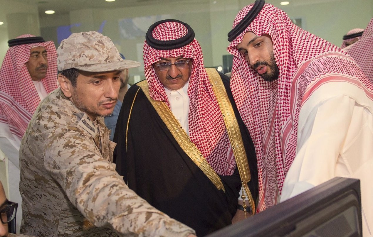 L'image montre un groupe de personnes, dont des hommes en tenue militaire et en vêtements traditionnels. L'un des hommes en uniforme semble expliquer quelque chose à ses collègues, qui sont attentifs à un écran d'ordinateur. L'environnement semble être un bureau ou une salle de réunion, et l'atmosphère suggère une discussion sérieuse ou une présentation. Les hommes semblent de haut rang, probablement impliqués dans des affaires militaires ou gouvernementales.