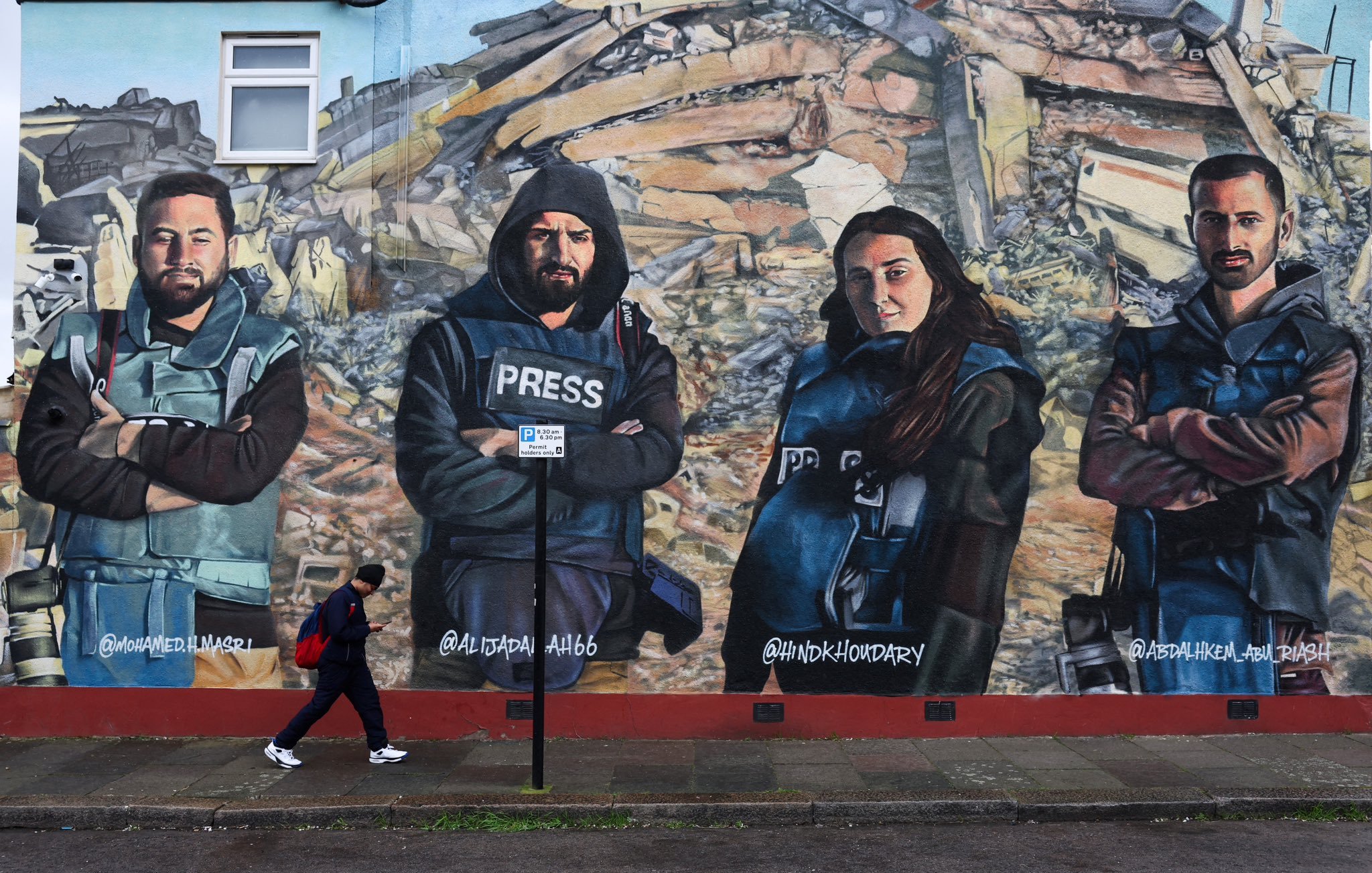 L'image montre une fresque murale représentant quatre personnes habillées en gilets pressés, posant avec les bras croisés. Ils semblent symboliser des journalistes ou des reporters. En arrière-plan, il y a des débris, suggérant un contexte de destruction ou de conflit. Une personne marche au premier plan, ajoutant une dynamique à la scène. Les visages des individus sur la fresque sont sérieux et déterminés.