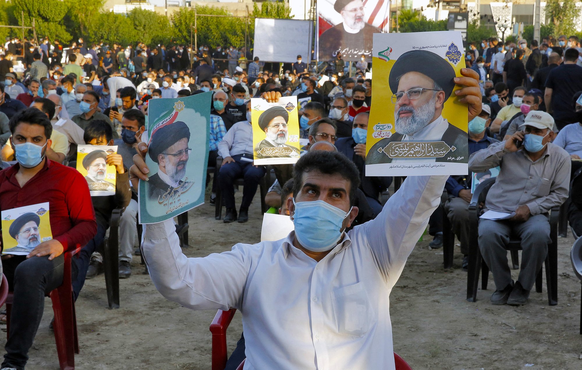L'image montre une assemblée de personnes rassemblées en plein air, assises sur des chaises. La plupart des participants portent des masques. Un homme, au premier plan, lève une affiche représentant une figure politique, probablement un leader ou un candidat, symbolisant un soutien. Les autres personnes autour de lui tiennent également des affiches similaires. En arrière-plan, on peut apercevoir des arbres et des structures, suggérant un rassemblement organisé. L'atmosphère semble être celle d'un événement politique ou d'une manifestation.