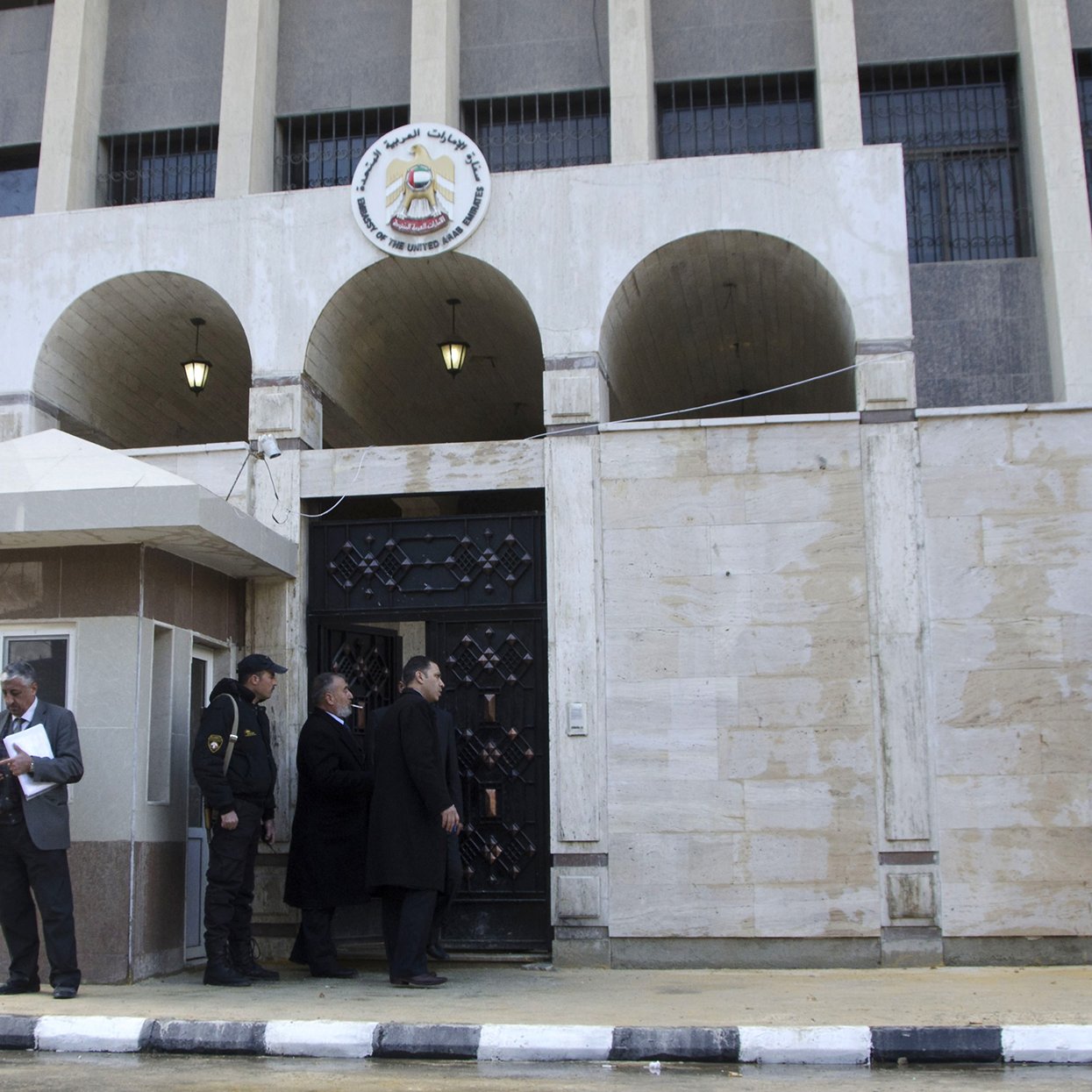 L'image montre un bâtiment arborant une façade en pierre. À l'avant, on voit une entrée avec des portes en fer forgé, encadrée par des arches. À gauche, un homme en costard consulte un document, tandis que deux autres hommes, également en costume, discutent à l'entrée. Un homme est sur une échelle, en train de fixer un panneau sur le mur. En arrière-plan, on distingue un poste de sécurité, suggérant que c'est un site administratif ou gouvernemental. Le sol est pavé en noir et blanc, ajoutant un contraste au décor.