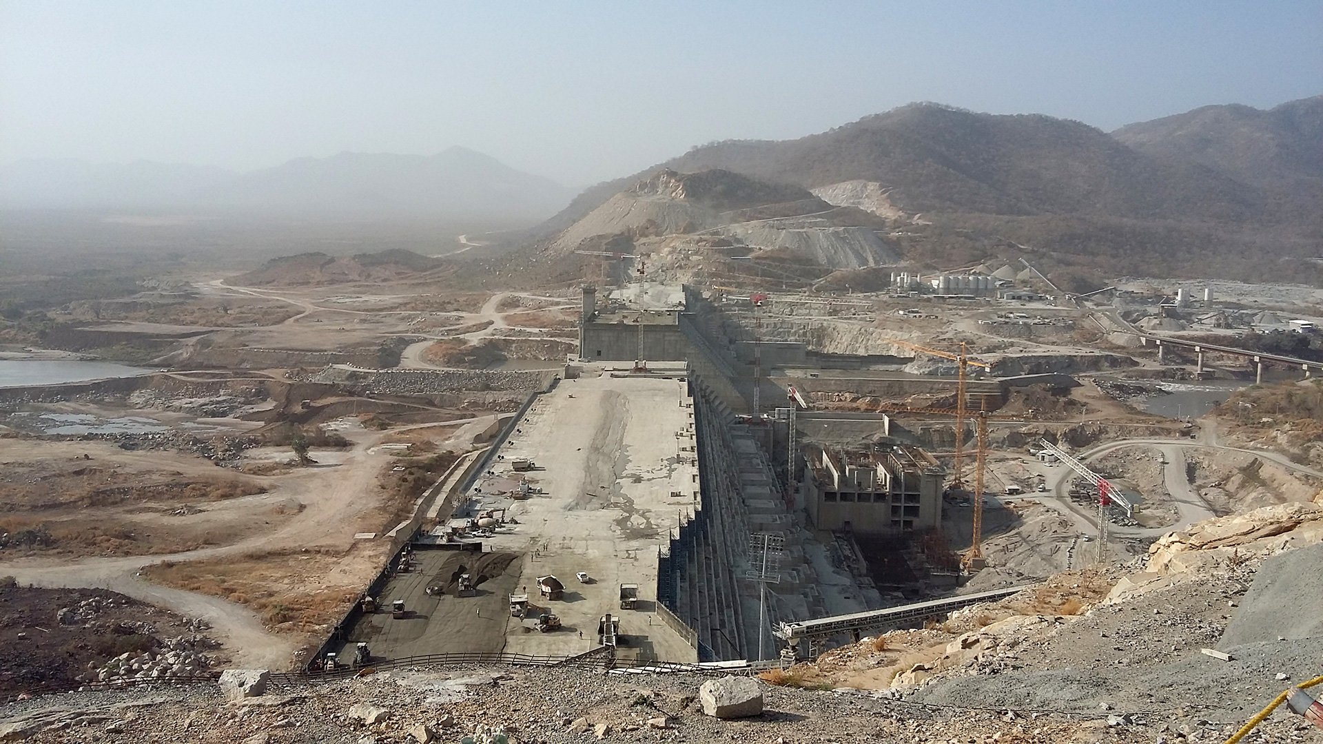 The image depicts a large, under-construction dam situated in a mountainous landscape. The dam site features extensive concrete structures and machinery, indicating significant engineering work in progress. Surrounding the site, there are vast areas of earth that have been moved or excavated, along with roads and pathways that suggest ongoing construction activity. The landscape is somewhat arid, with sparse vegetation and mountainous terrain in the background. The visibility is limited, possibly due to haze or dust in the atmosphere.