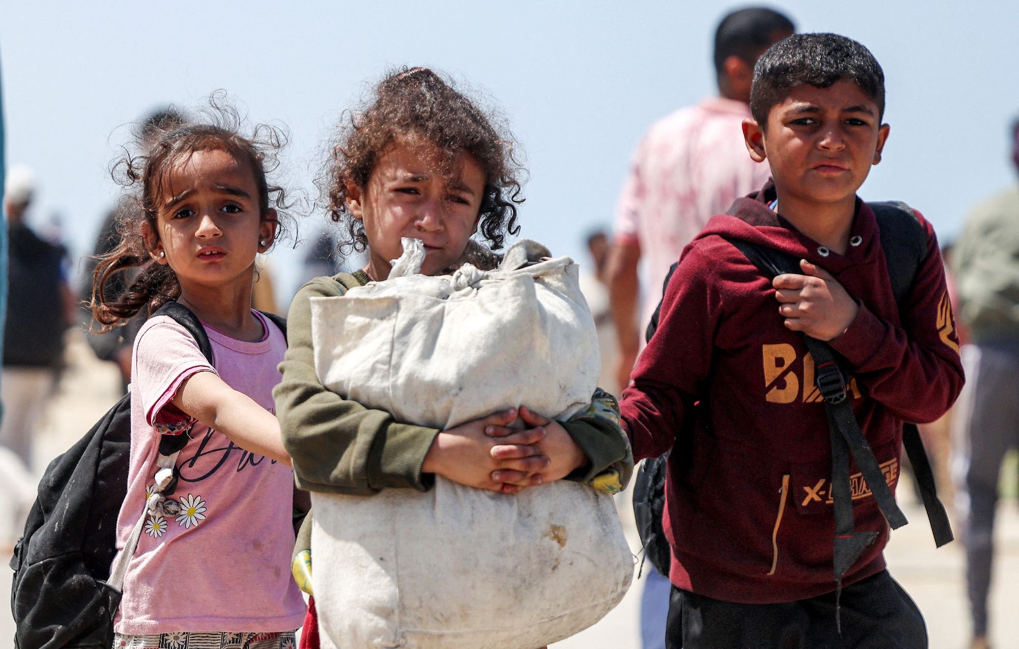 L'image montre un groupe d'enfants se déplaçant ensemble. Deux jeunes filles portent des vêtements simples, l'une d'elles a les cheveux bouclés et tient un grand sac. Le garçon à droite, portant un sweat à capuche rouge, semble également porter un sac. Ils expriment une certaine détresse, et l'arrière-plan suggère un environnement de crise ou de déplacement. La scène évoque des thèmes de vulnérabilité et de résilience.